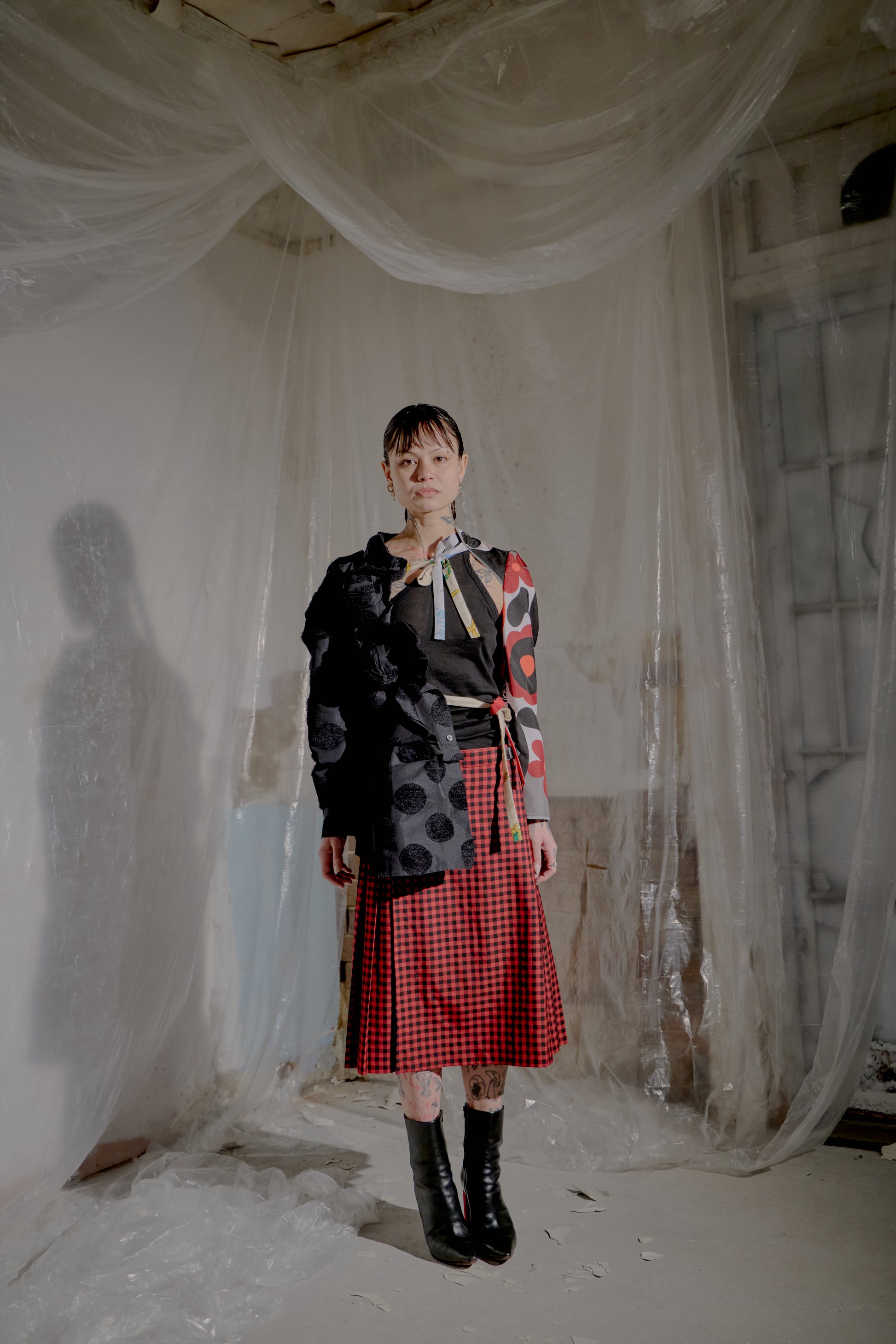 Person wearing a black Comme Des Garcons jacket with colourful patterns and a red plaid skirt in a room with white curtains.