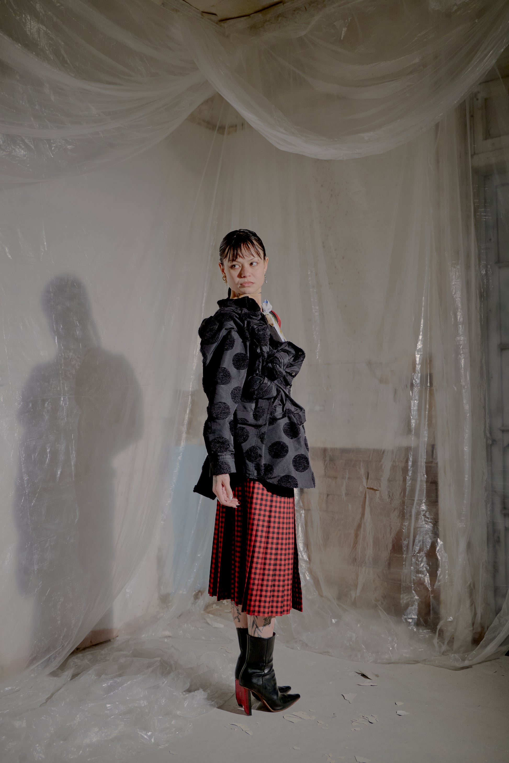 Person wearing a black polka dot Comme Des Garcons jacket and red checkered skirt standing in a room with plastic sheeting.