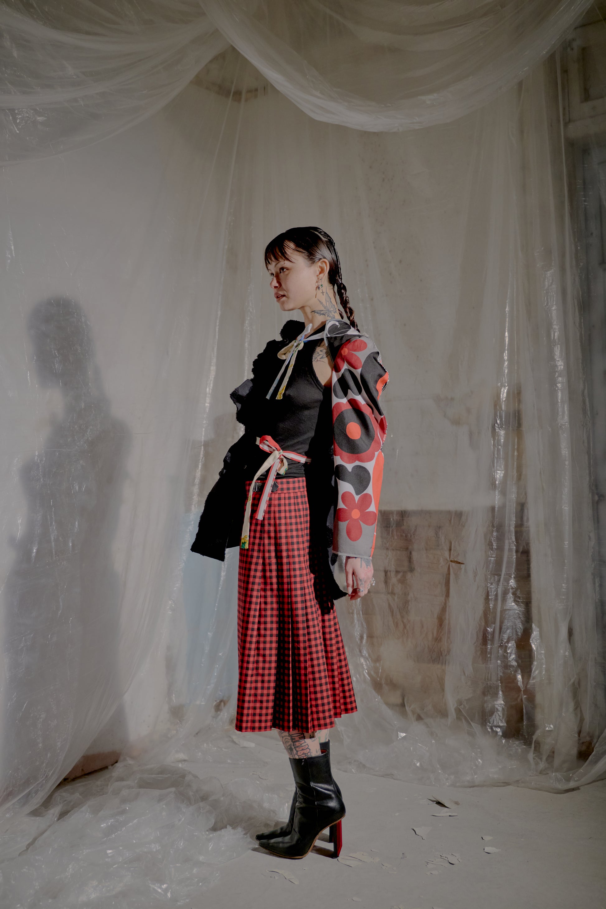 Person wearing a black Comme Des Garcons, red plaid skirt, and black boots in a draped white fabric setting.