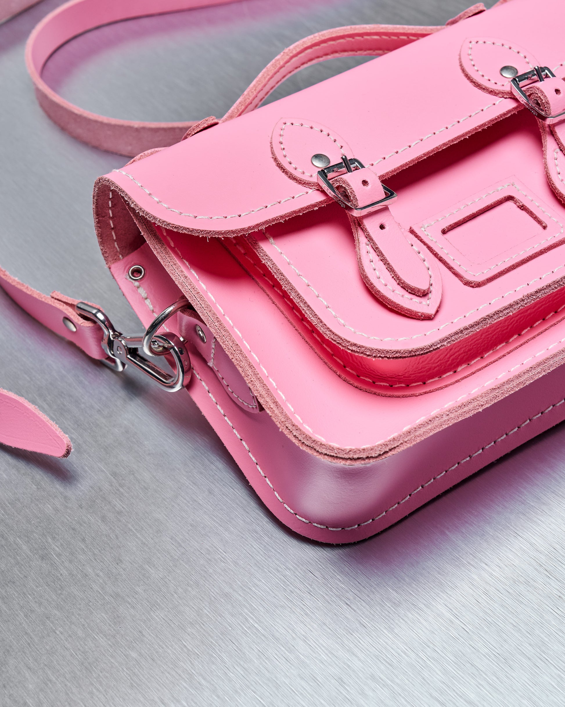 Close-up of a vibrant pink leather Comme des Garcons satchel on a metallic surface. The bag features buckled straps, contrast stitching, and a small handle, conveying a chic and playful tone.