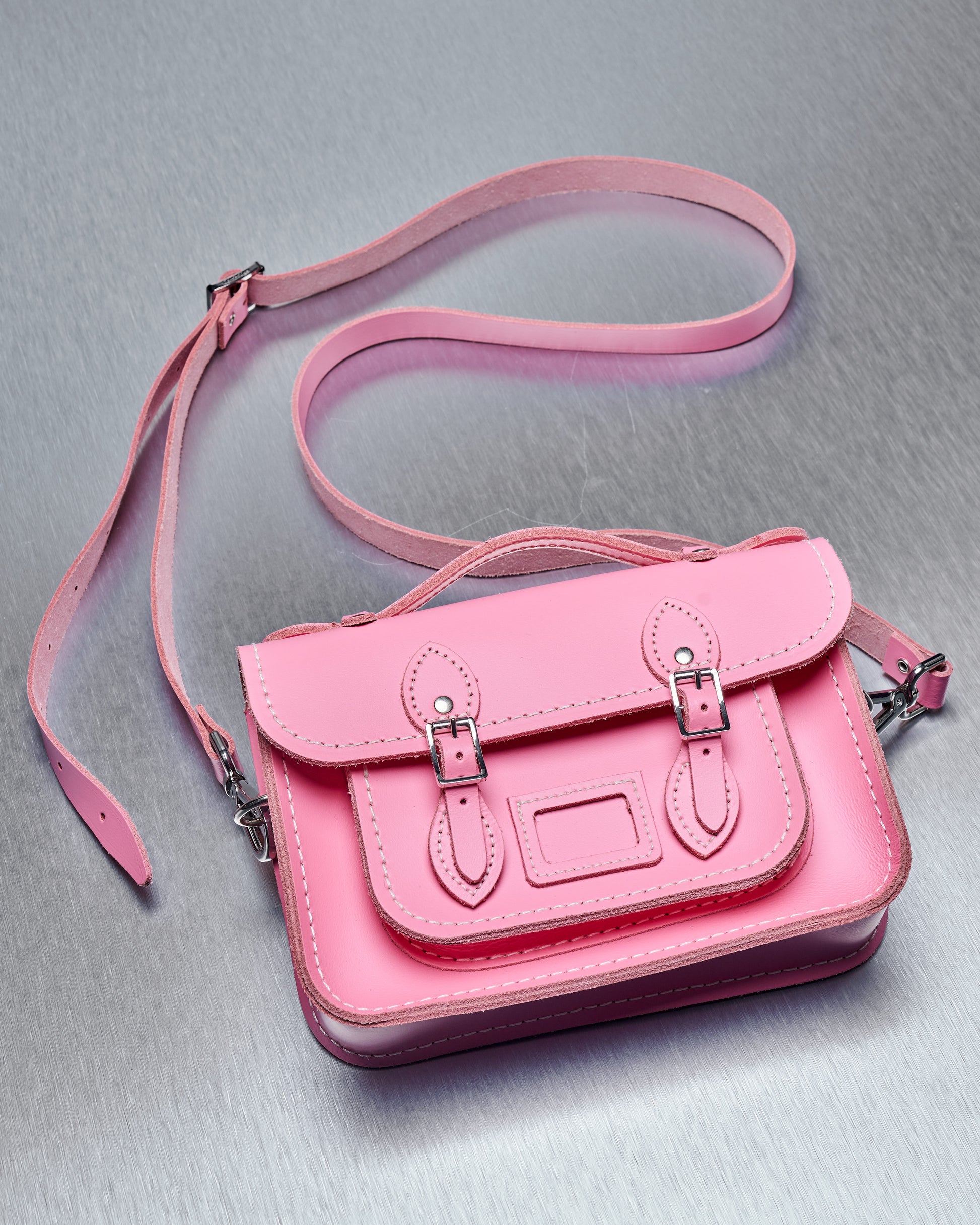 Close-up of a vibrant pink leather Comme des Garcons satchel on a metallic surface. The bag features buckled straps, contrast stitching, and a small handle