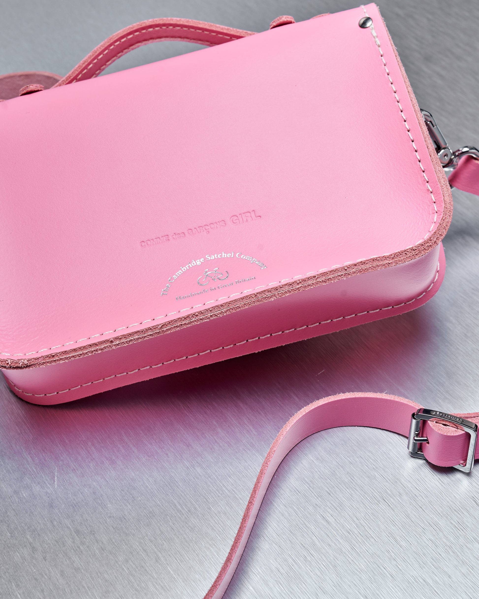 A pink leather Comme des Garcons satchel with a top handle and adjustable strap rests on a metallic surface. The bag has embossed branding, conveying a stylish tone.