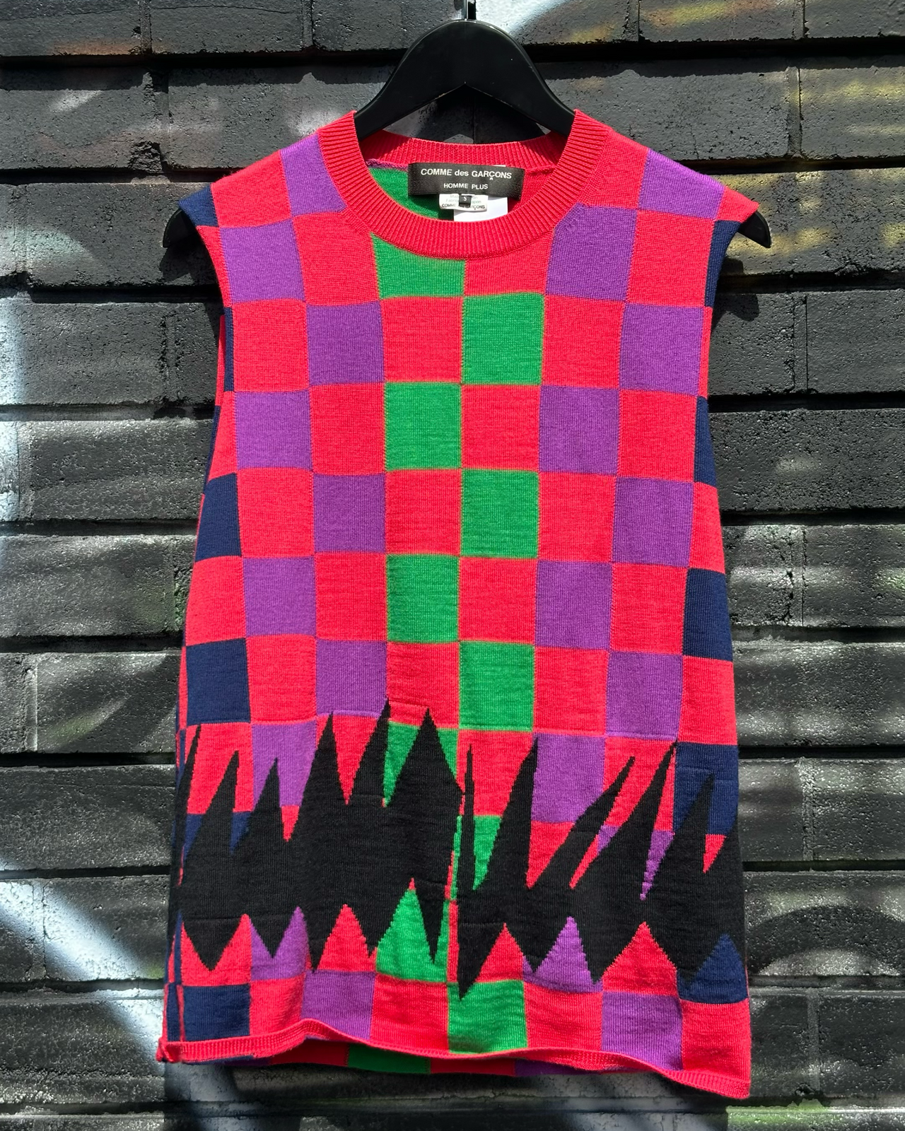 Colourful checkered sleeveless Comme des Garcons vest with geometric patterns on a textured wall background