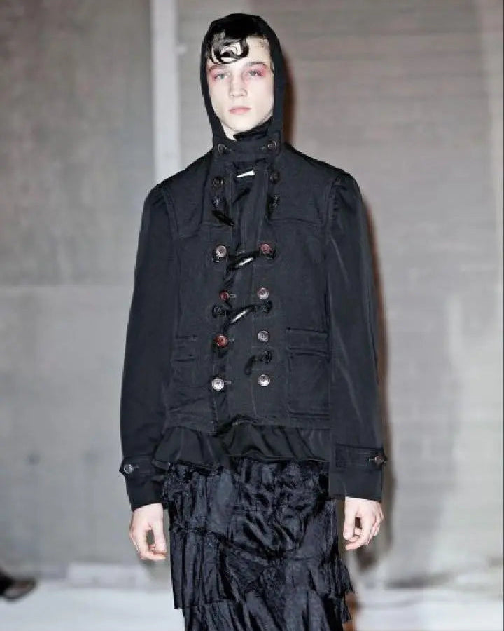Person wearing a dark, layered Comme Des Garcons outfit with a hood in an indoor setting