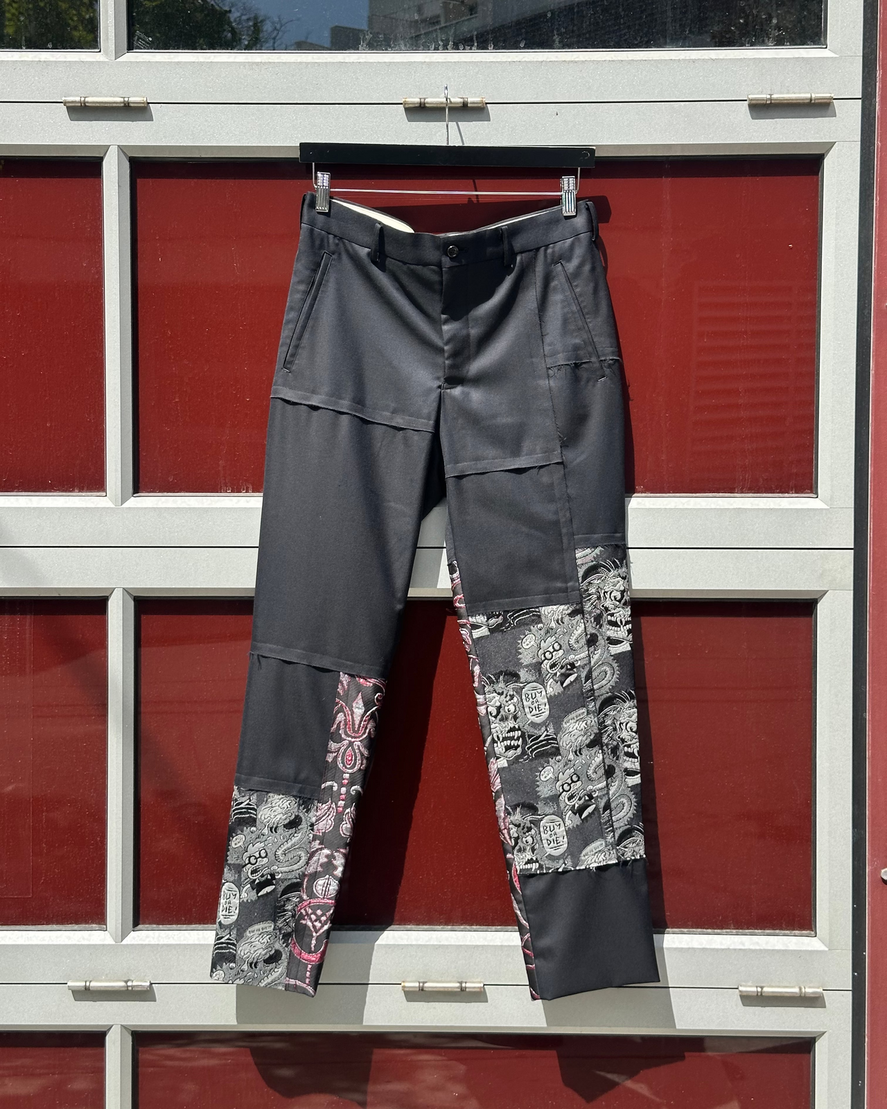 Dark gray Comme des Garcons pants with floral patterns hanging against a red and white building.