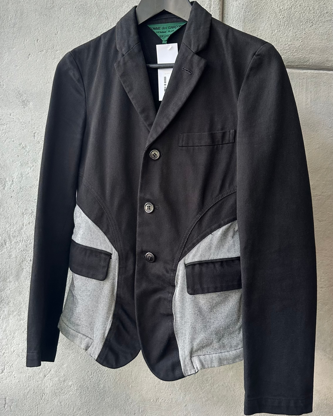 Black Comme des Garcons Homme Plus jacket with gray patchwork design on a hanger against a light gray background