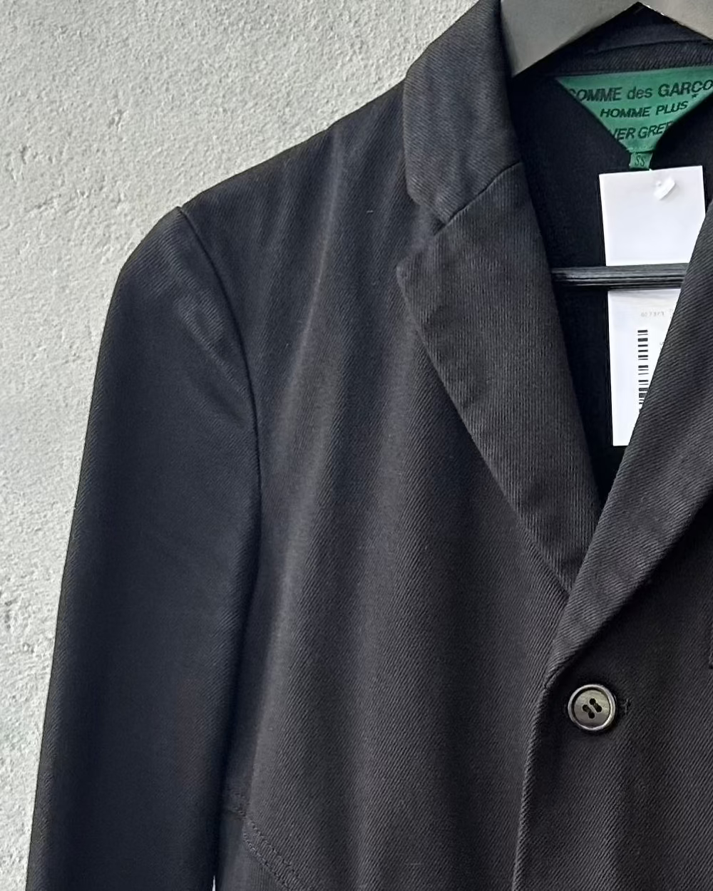 Dark gray Comme des Garcons blazer on a hanger with a visible brand tag against a light gray background