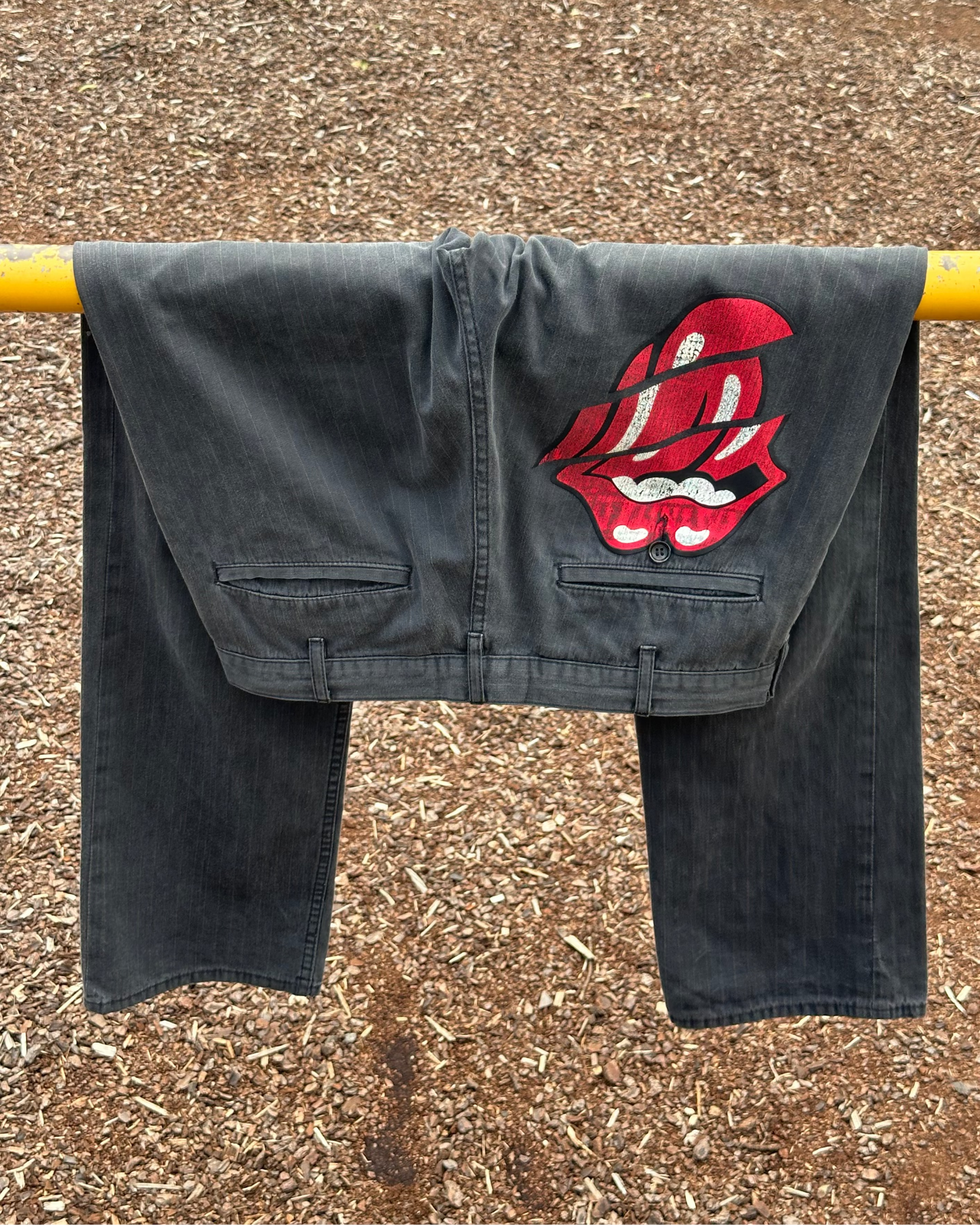 Black Comme des Garcons pants with a red and white design on a yellow rod against a brown ground.