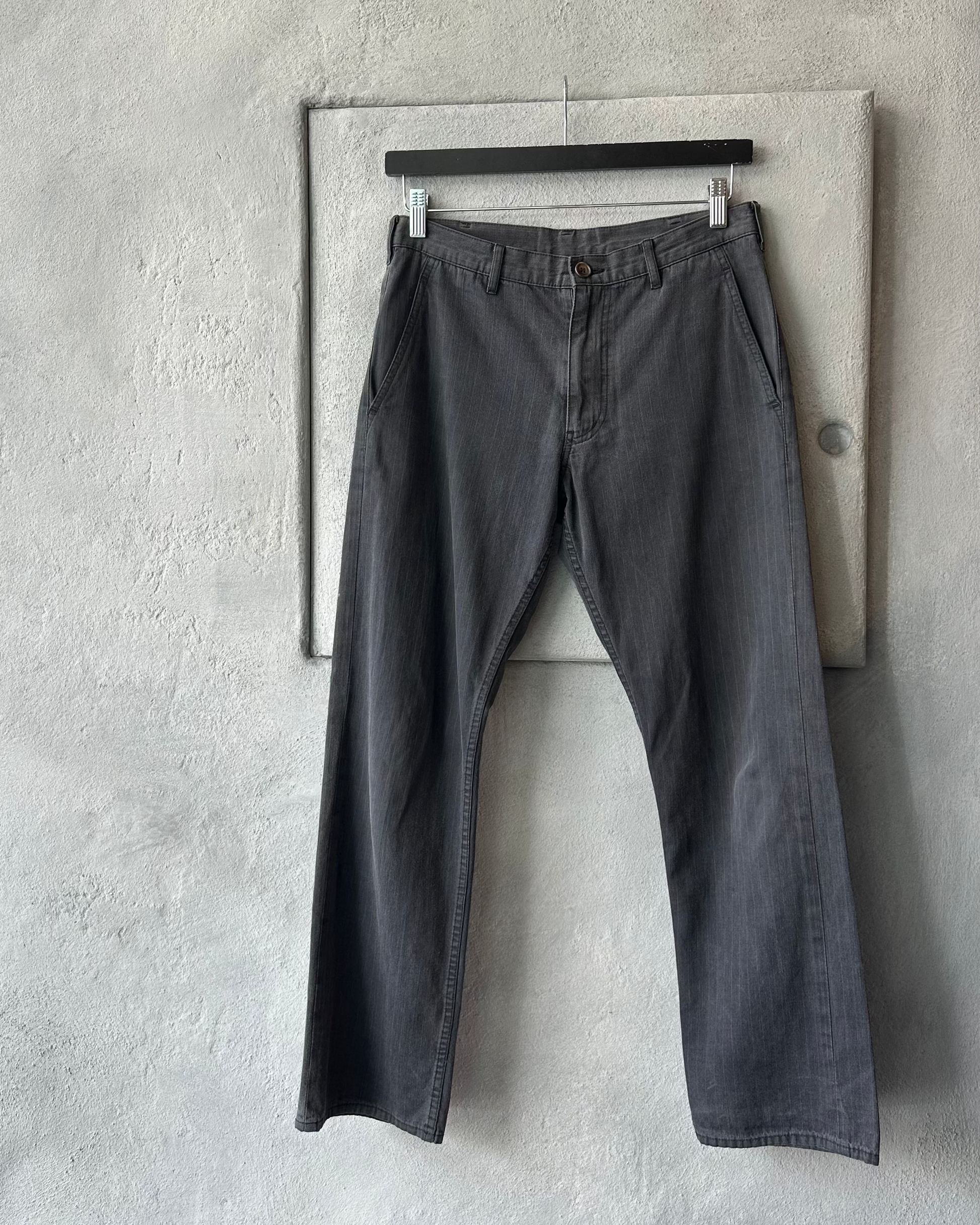 Pair of dark gray Comme des Garcons pants hanging on a wooden hanger against a light gray concrete wall.