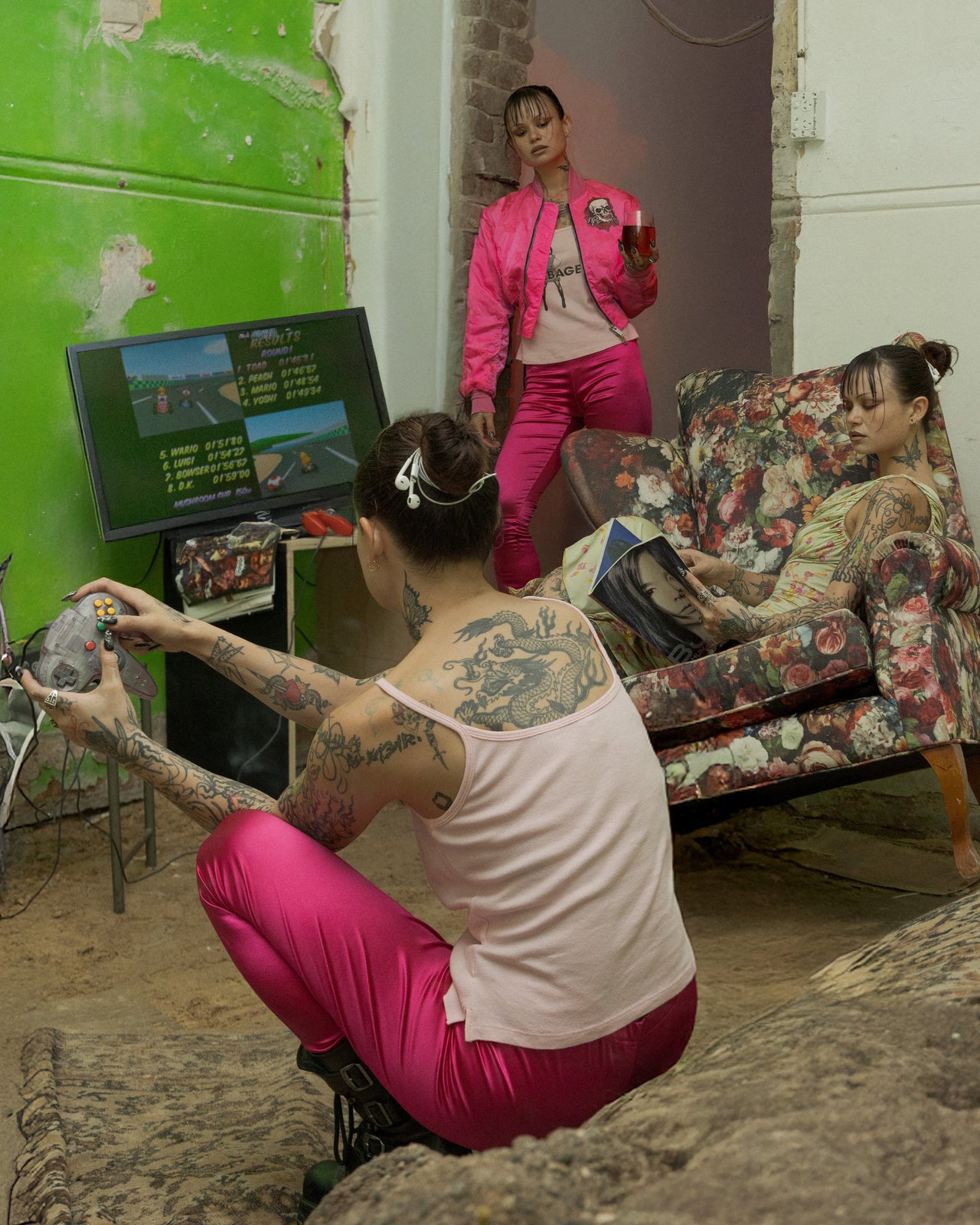 Three people in a room with a green wall, arcade machine, and floral couch.
