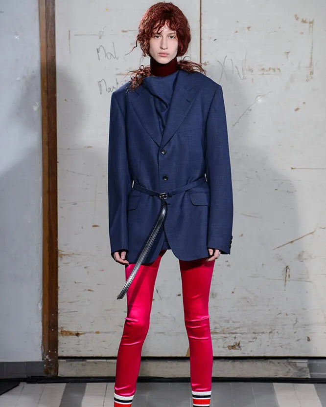 Person wearing a blue Comme des Garcons Junya Watanabe blazer and red pants standing against a textured wall.