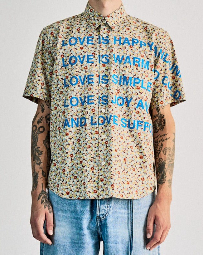 A person is standing, wearing a floral Comme des Garcons Junya Watanabe collared shirt with a glossy texture on the chest and sides, paired with blue jeans and white sneakers.