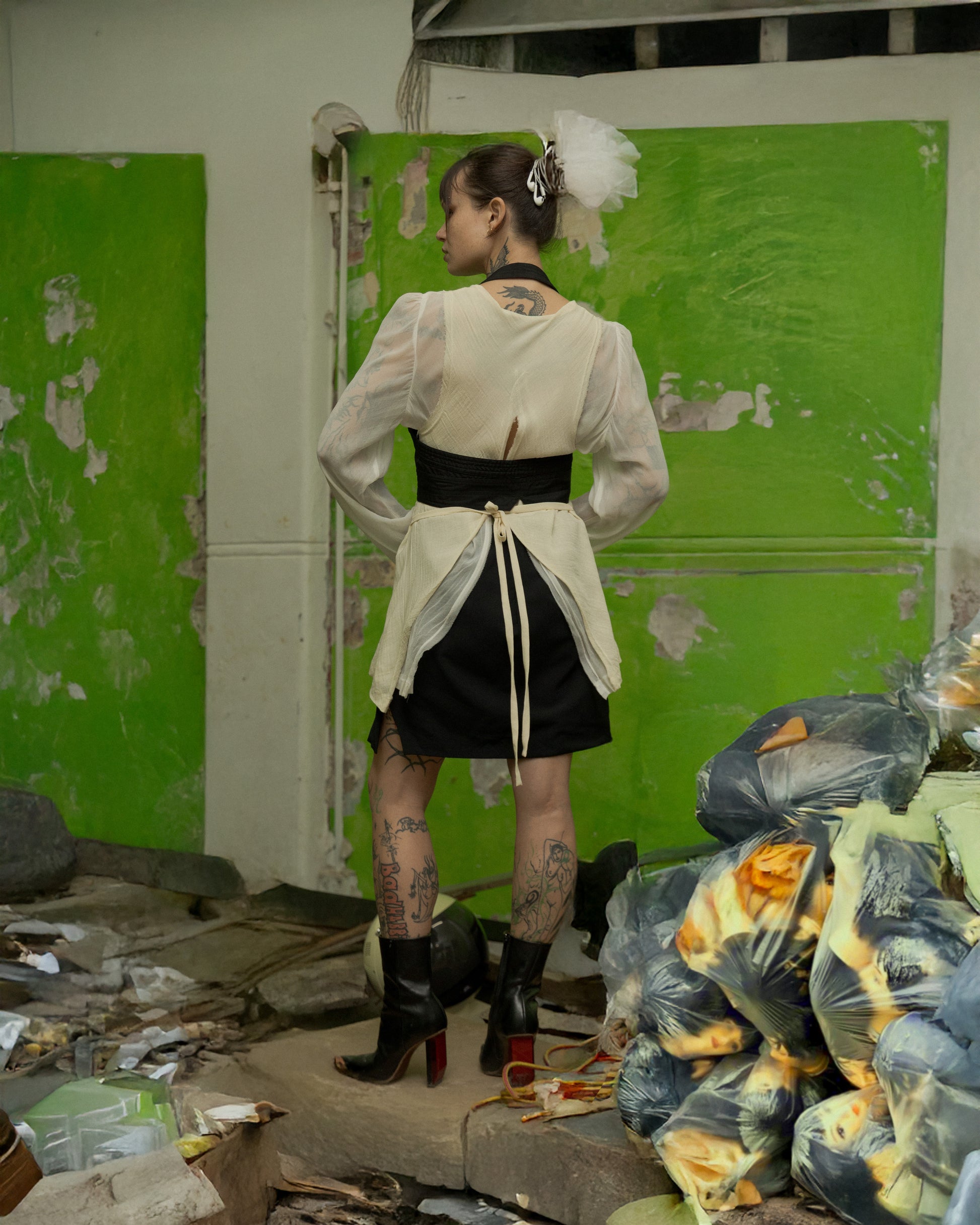Person wearing a white Comme Des Garcons blouse and black skirt in a dilapidated room with green walls and debris.