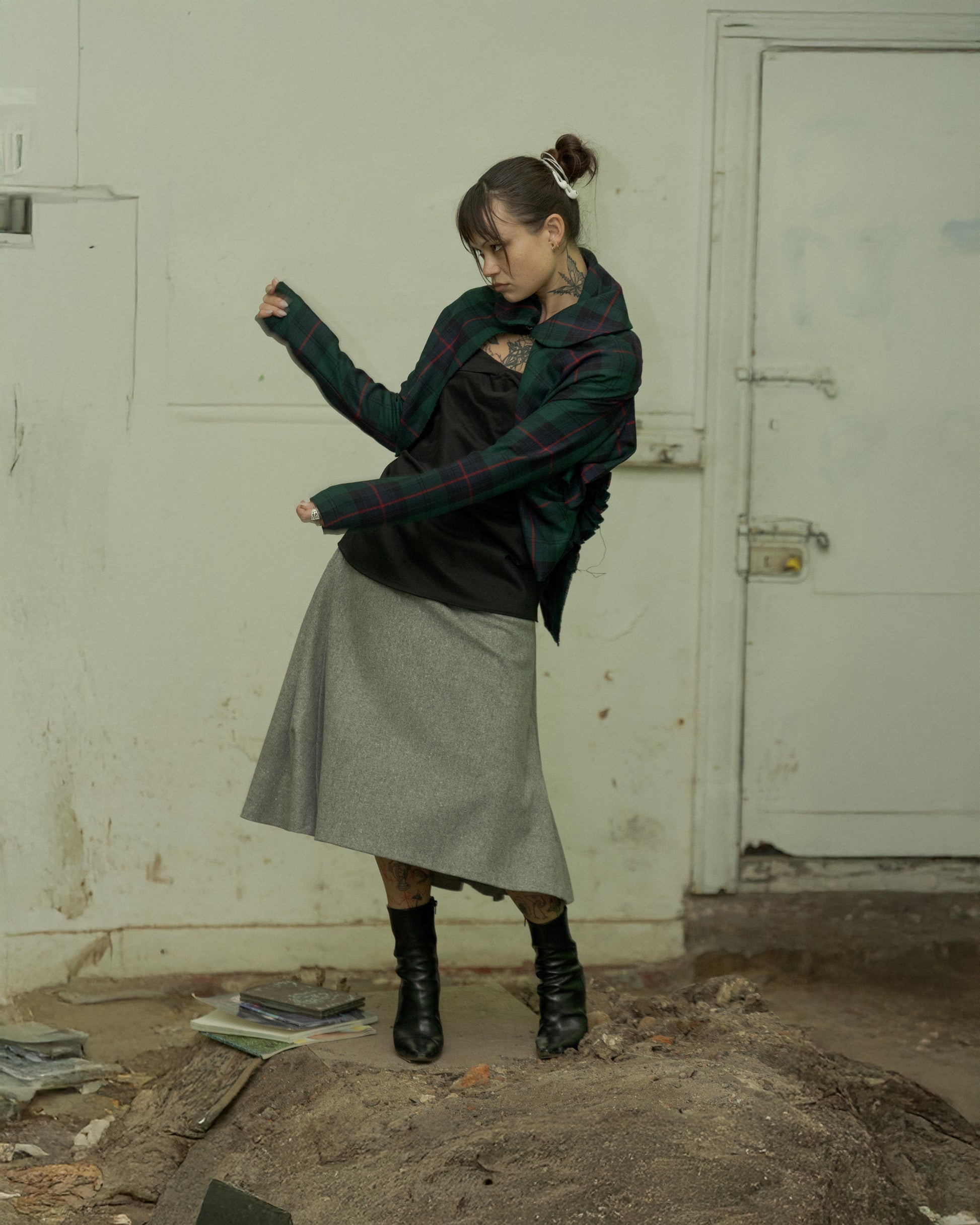 A person wearing a Comme des Garçons tartan asymmetrical jacket with an asymmetrical skirt and black boots, standing in a dilapidated indoor setting.
