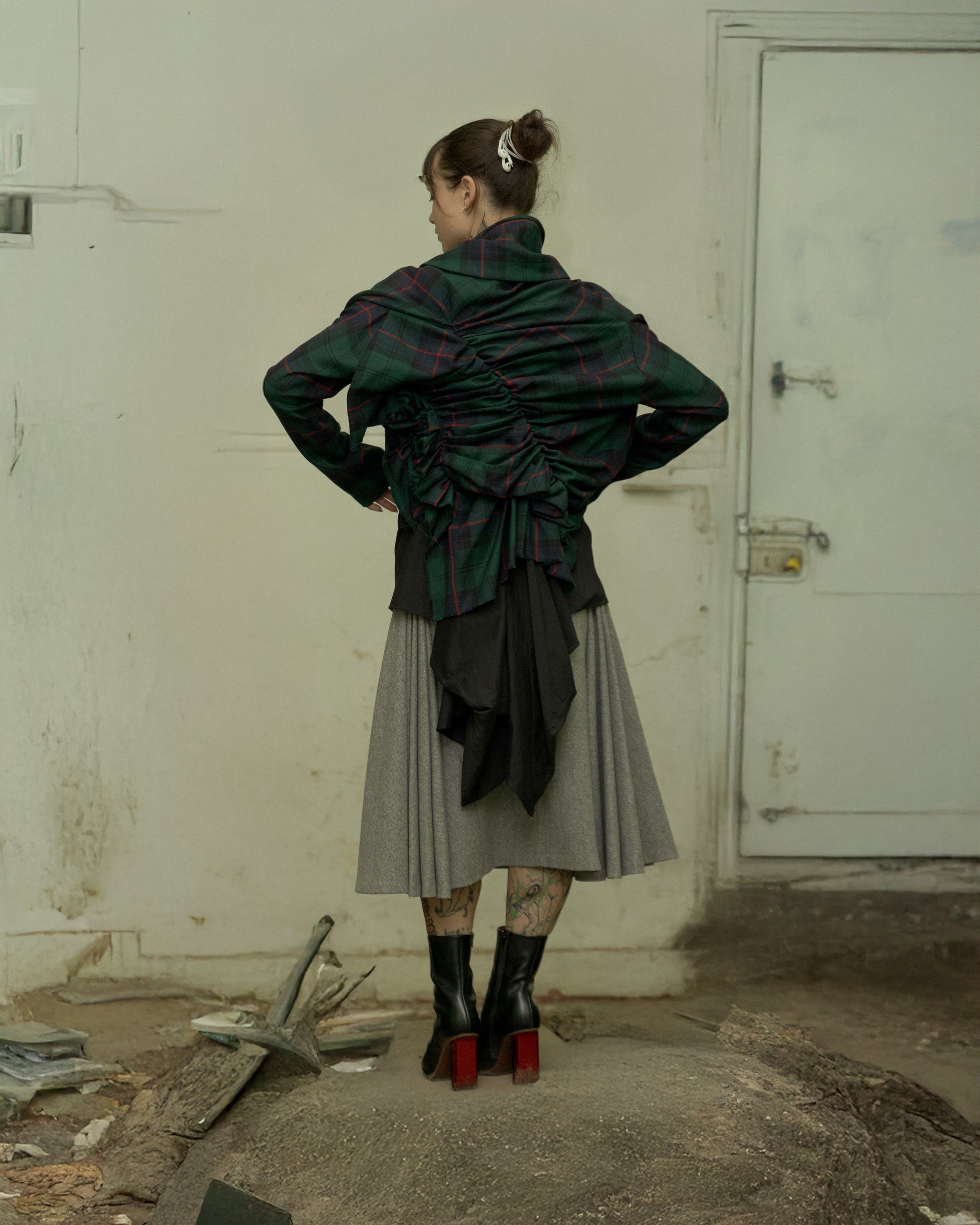 Person wearing a dark green and black Comme Des Garcons jacket with a gray skirt in an industrial setting.