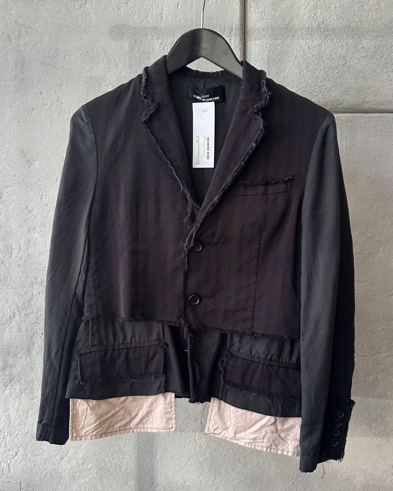 Black Comme des Garcons jacket with a beige lining on a hanger against a gray wall.