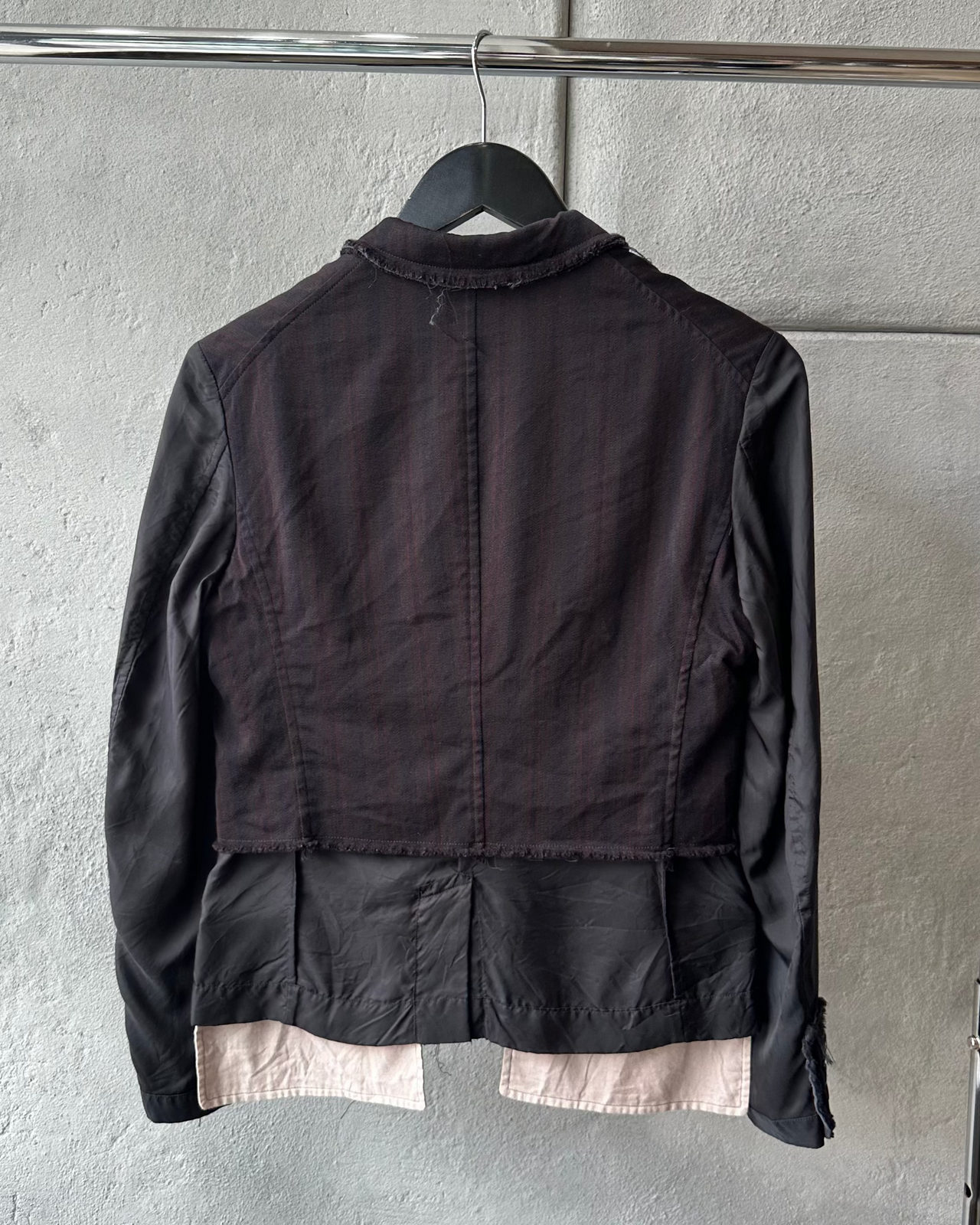 Black Comme des Garcons jacket with a beige lining on a hanger against a gray wall.