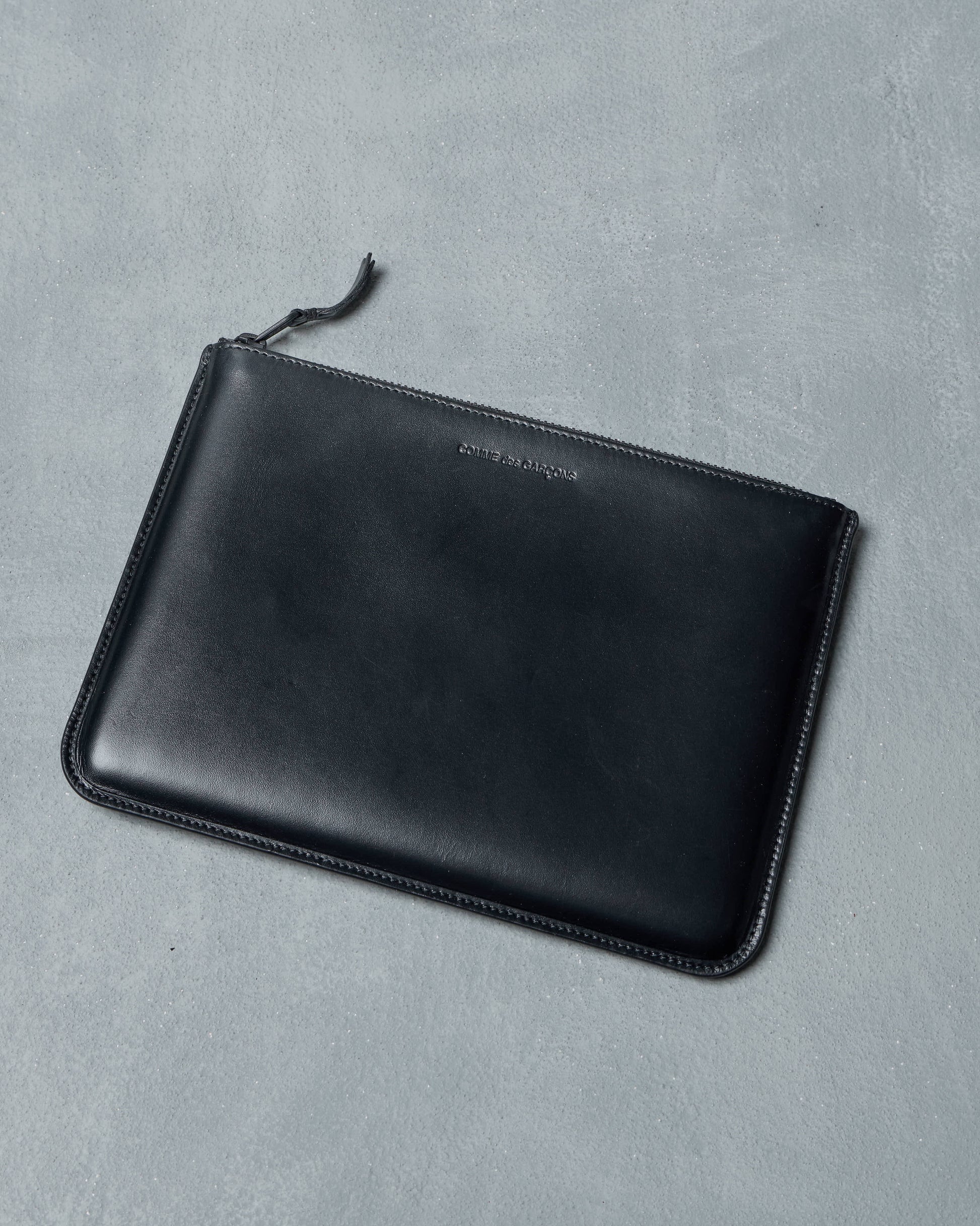 Black leather Comme des Garcons pouch with brand name on a gray background