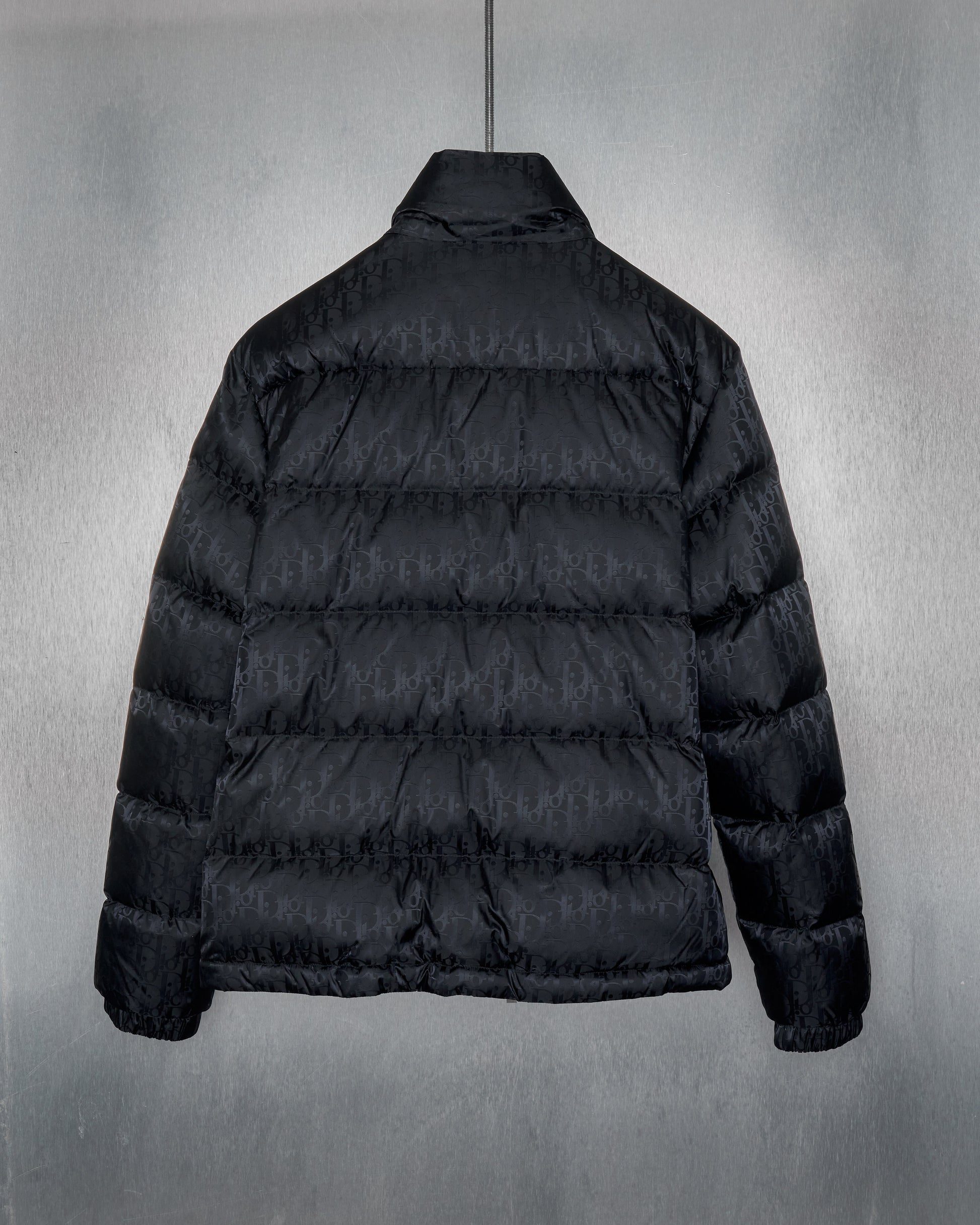 Black Dior puffer jacket with subtle embossed pattern, displayed against a metallic textured background
