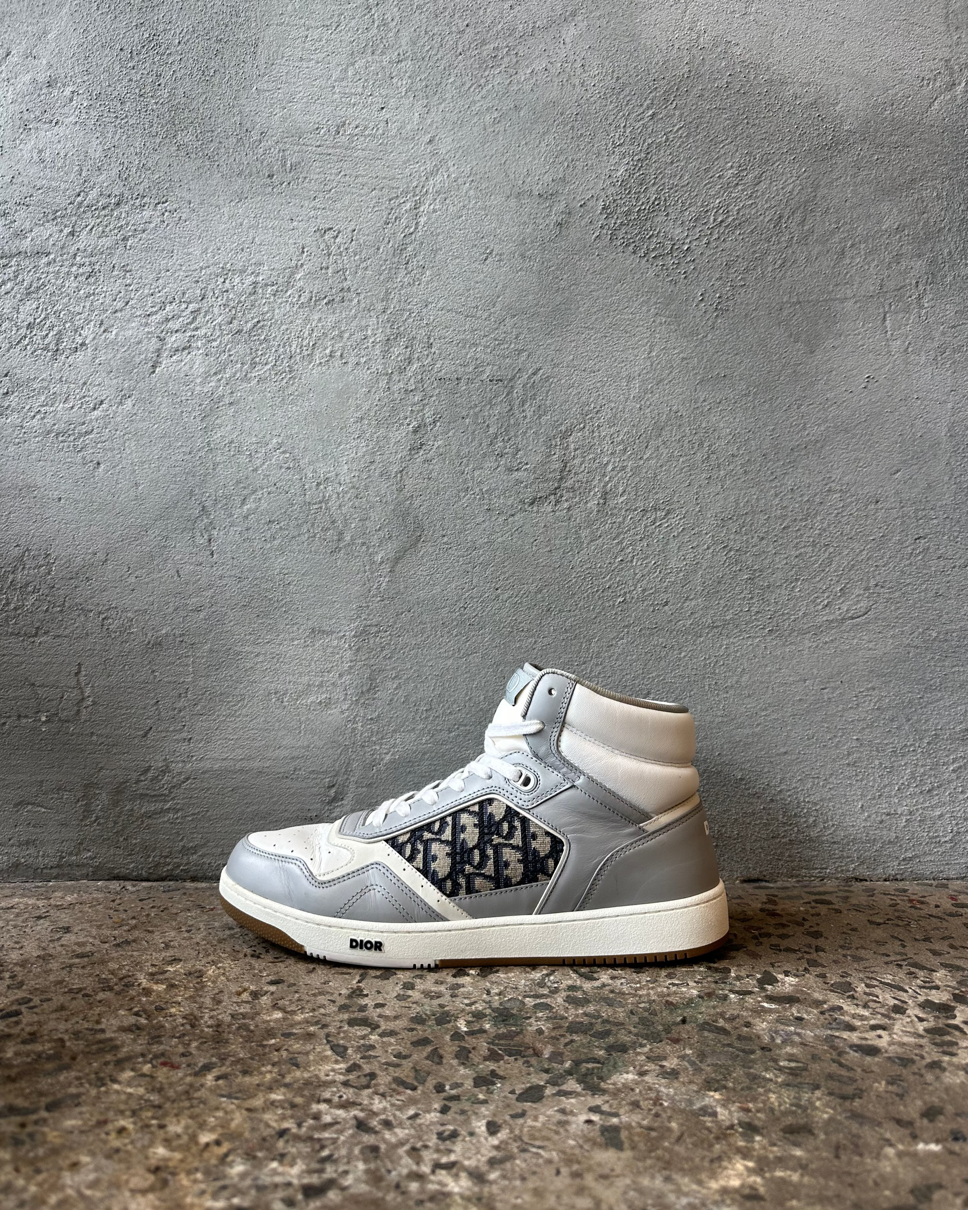 Gray high-top Dior sneaker with black pattern on a gray concrete surface and background