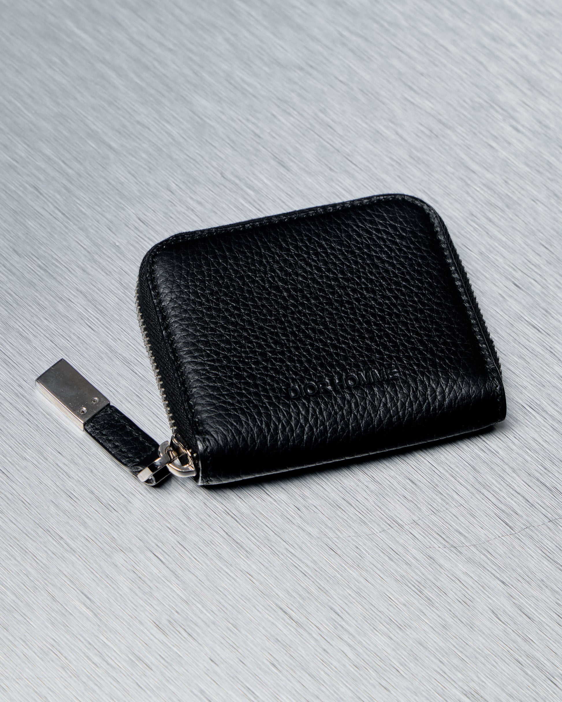 Black leather Dior Homme wallet with a zipper on a gray textured background