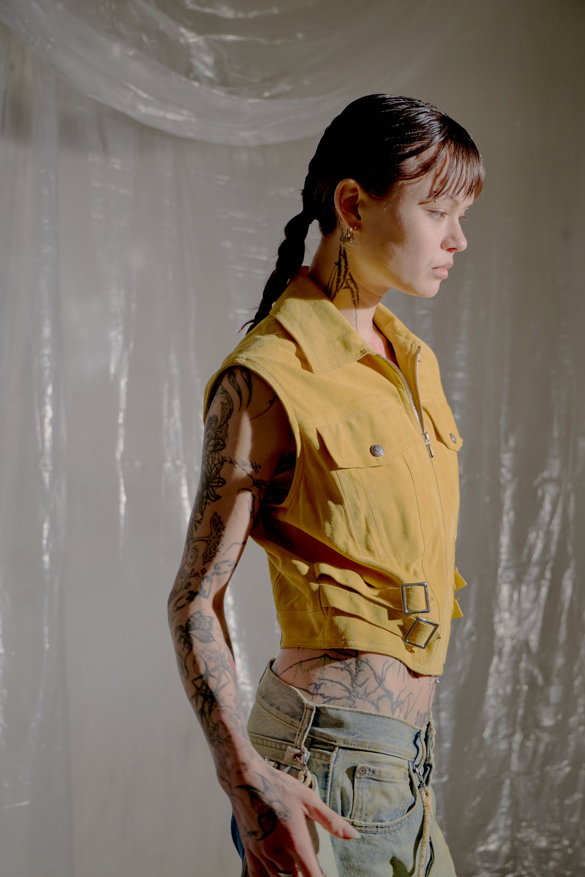 Person wearing a yellow Dior vest and light blue jeans with tattoos, standing against a plain background.