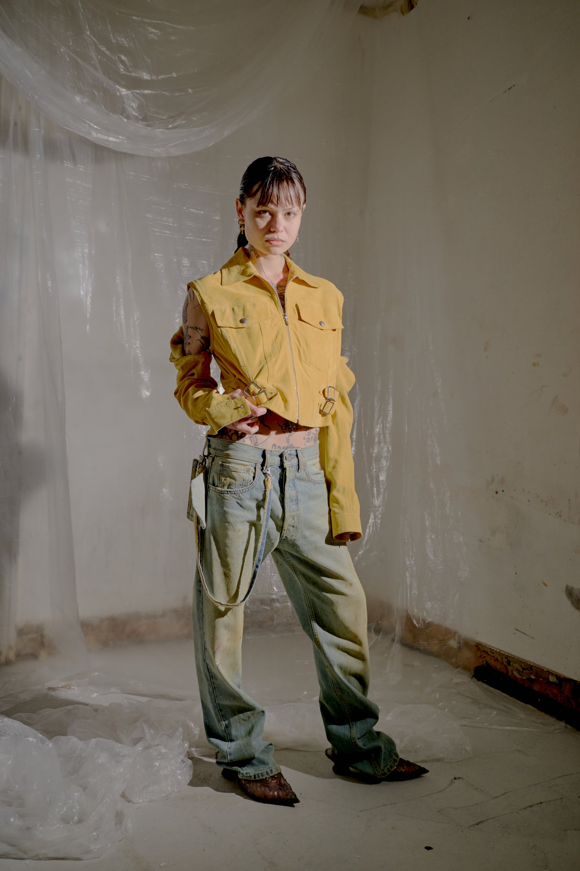 Person wearing a yellow Dior jacket and blue pants standing in a room with white walls and floor.