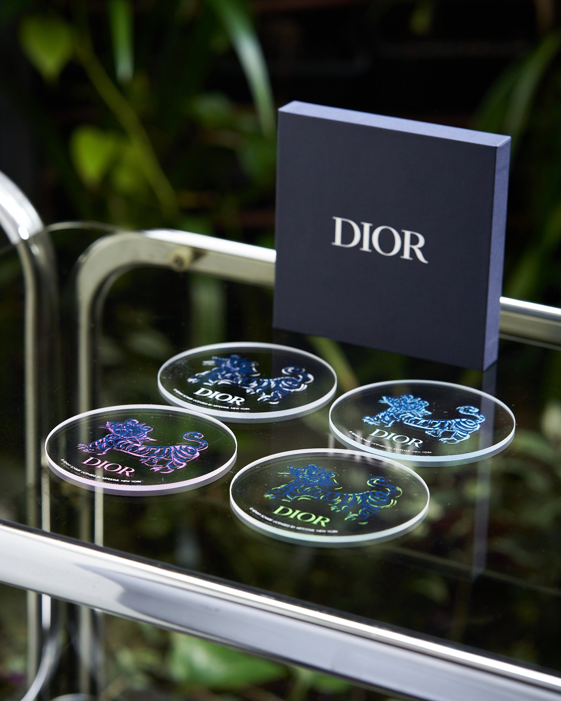 Circular coasters with colorful animal designs are arranged on a glass surface, next to a Dior-branded box. The setting is elegant and modern.
