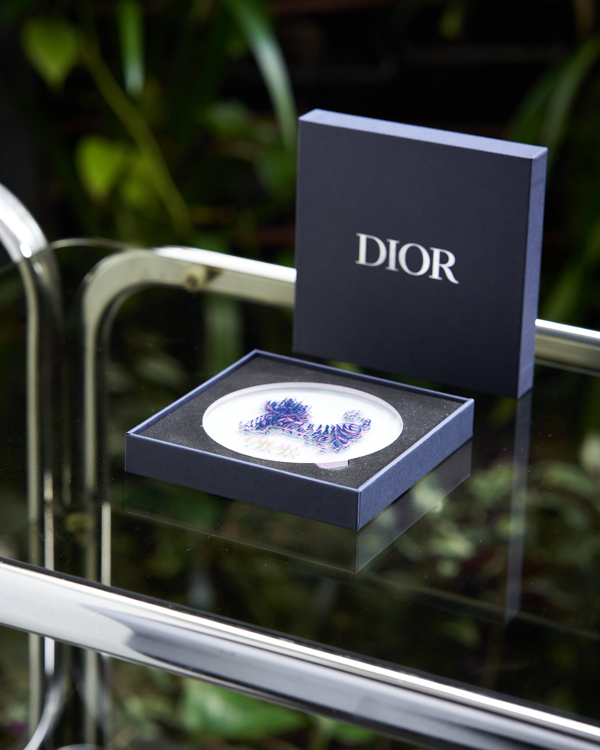 A blue Dior box on a reflective glass table contains an intricately designed brooch with blue and purple hues. 