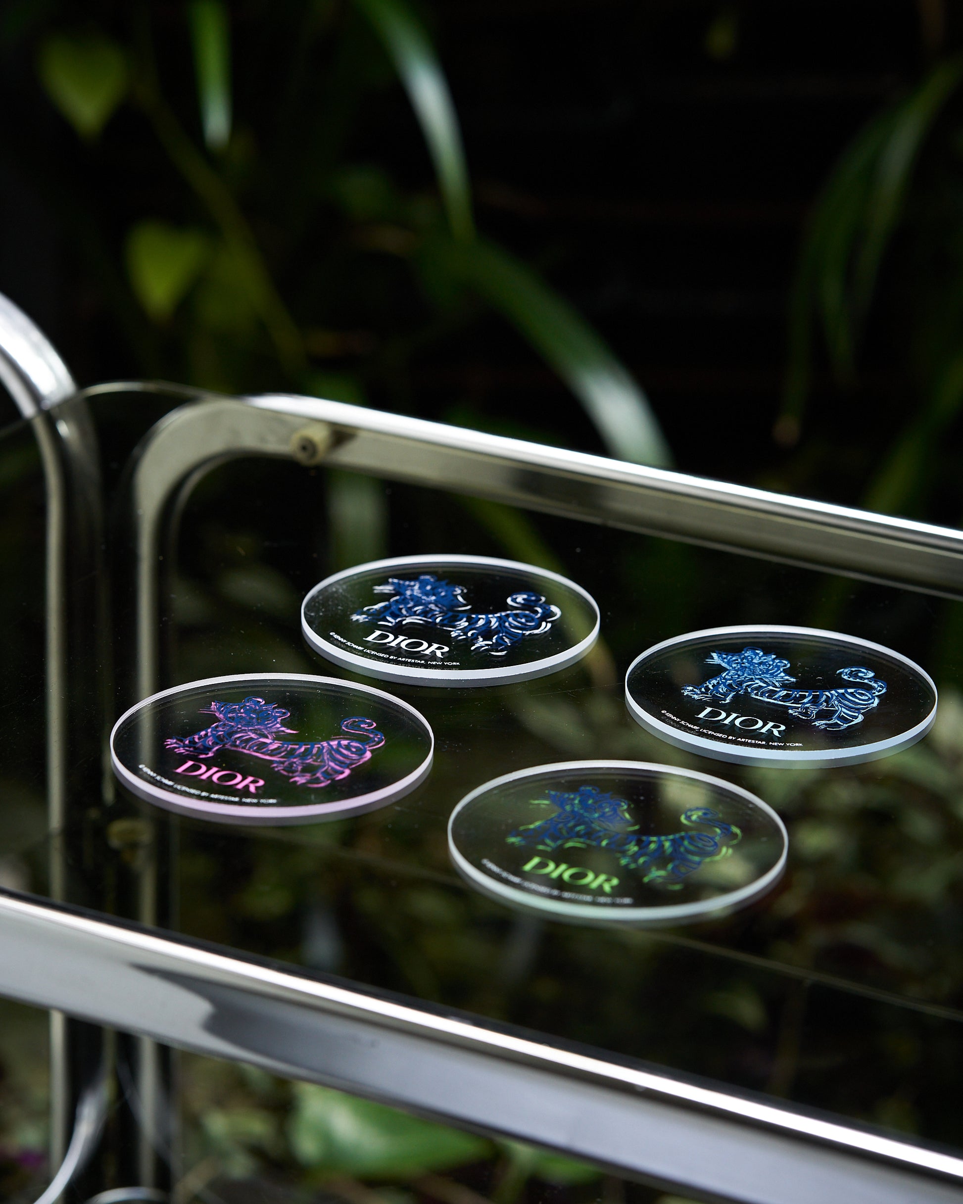 Four round coasters with colorful tiger designs and the "Dior" logo are displayed on a reflective glass surface. Green foliage is blurred in the background.