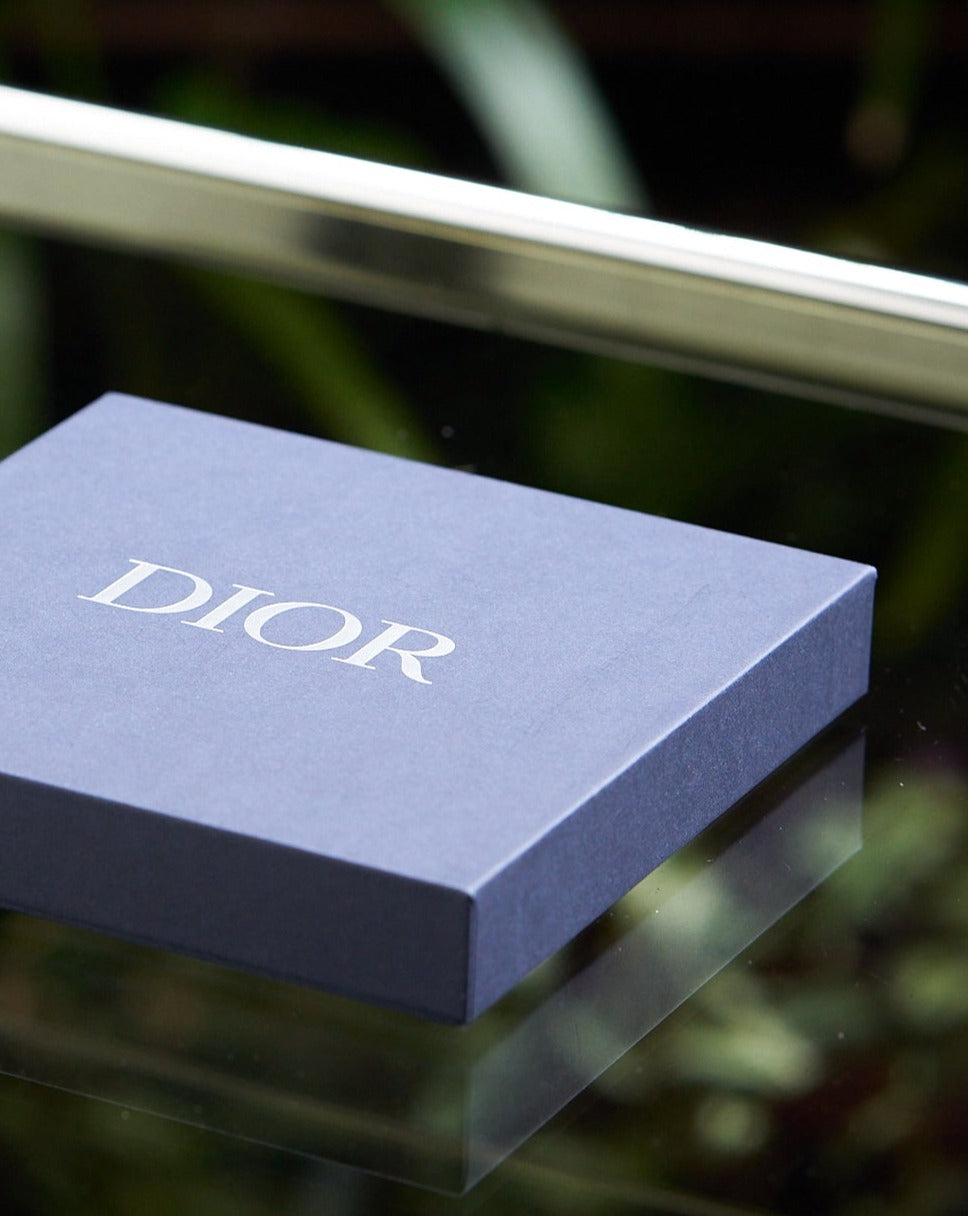 A dark blue Dior box rests on a reflective glass surface. The box is elegantly simple, conveying luxury and sophistication. Blurred green foliage is in the background.