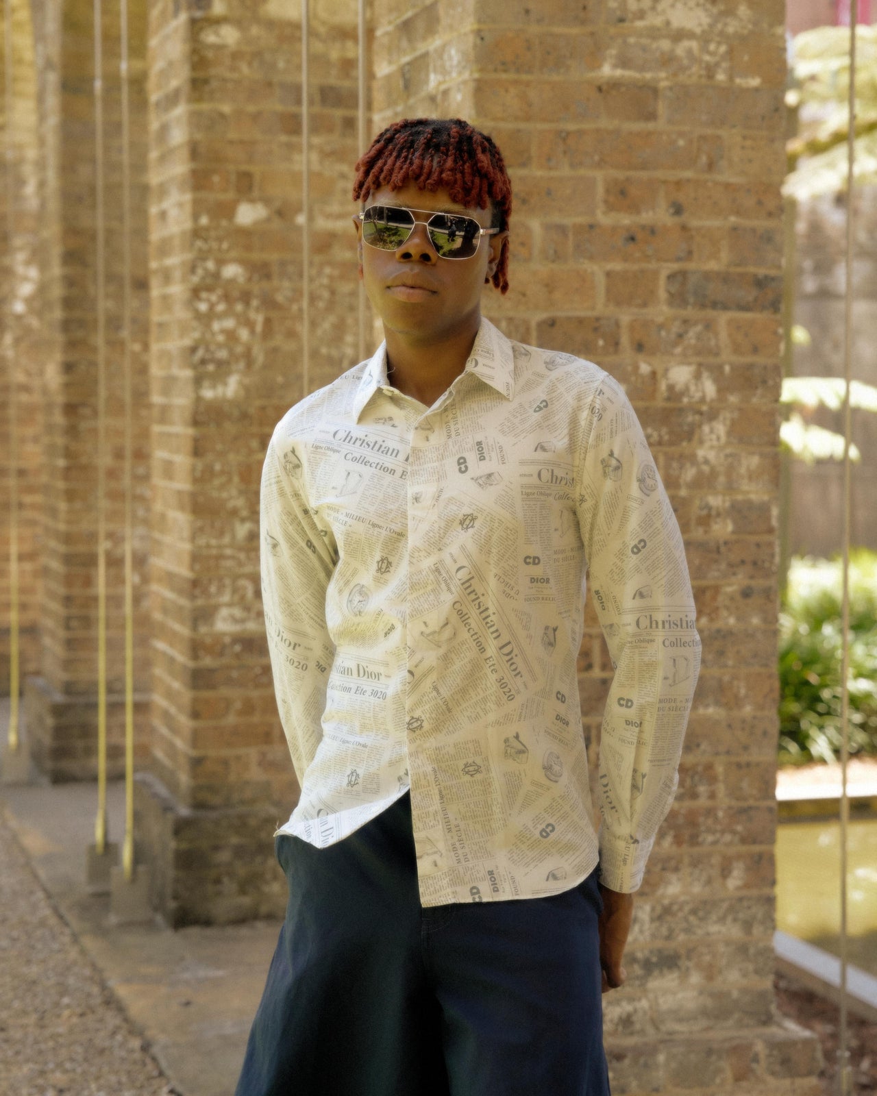 A person wearing sunglasses and a collared shirt with an all-over print of text in a Dior x Daniel Arsham SS 2020 style.