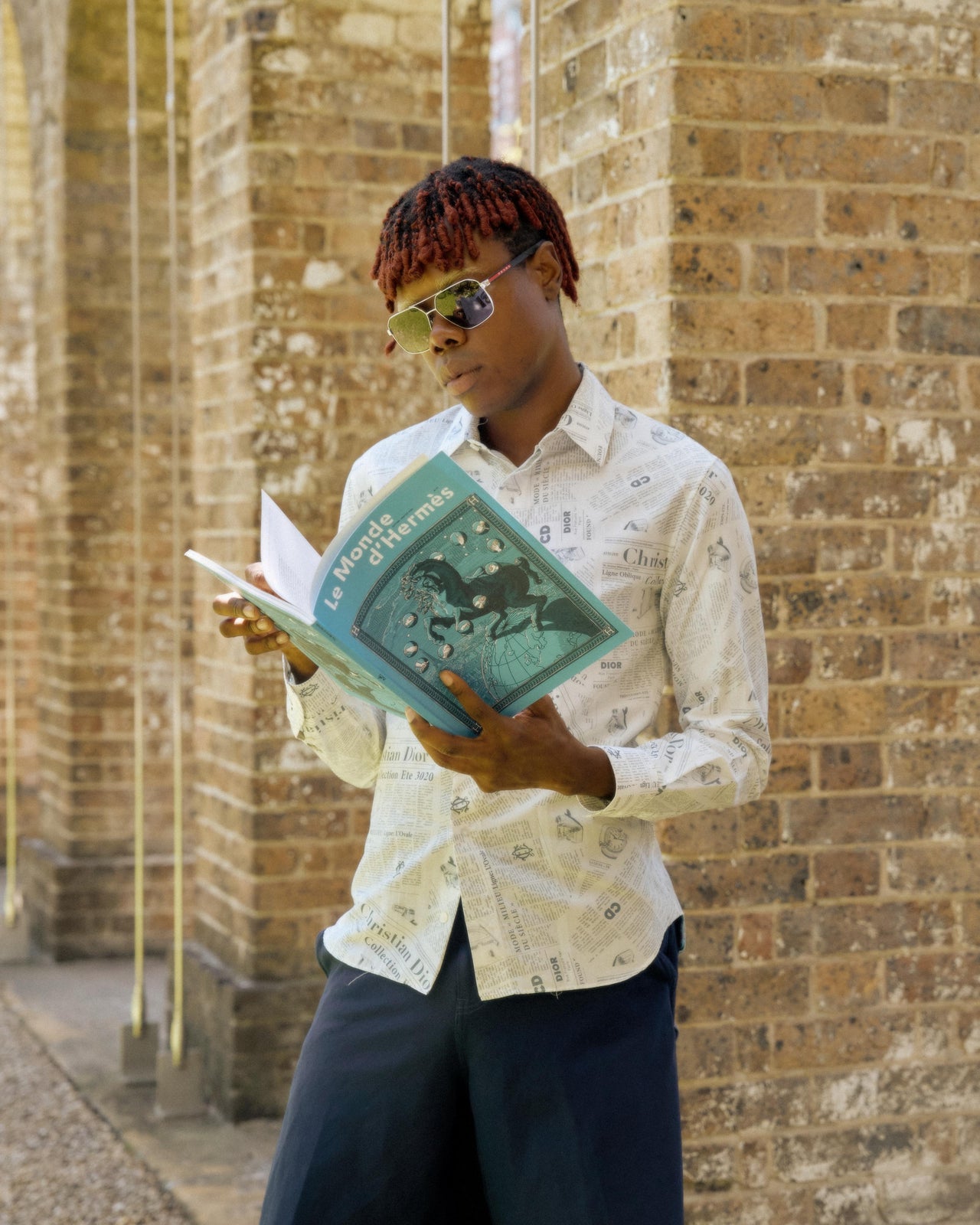 A person wearing sunglasses and a collared shirt with an all-over print of text in a Dior x Daniel Arsham SS 2020 style.