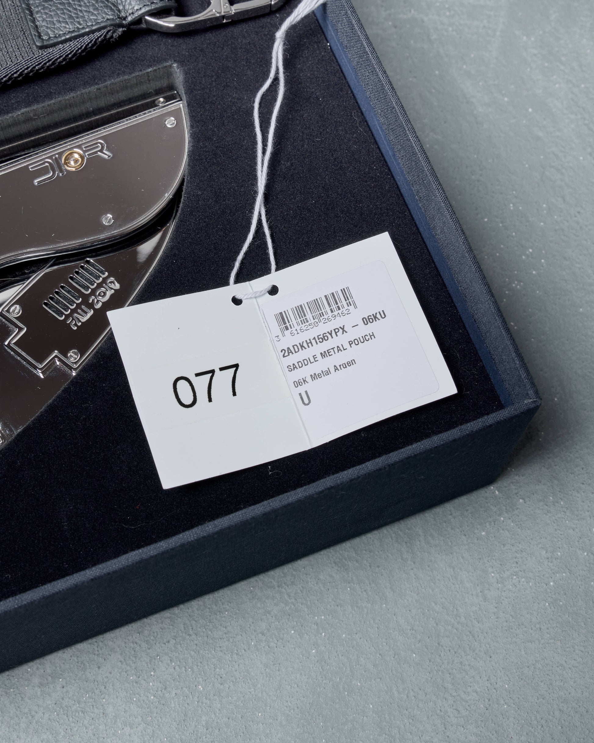 Silver Dior clutch packaging with a tag displaying '077' on a gray surface