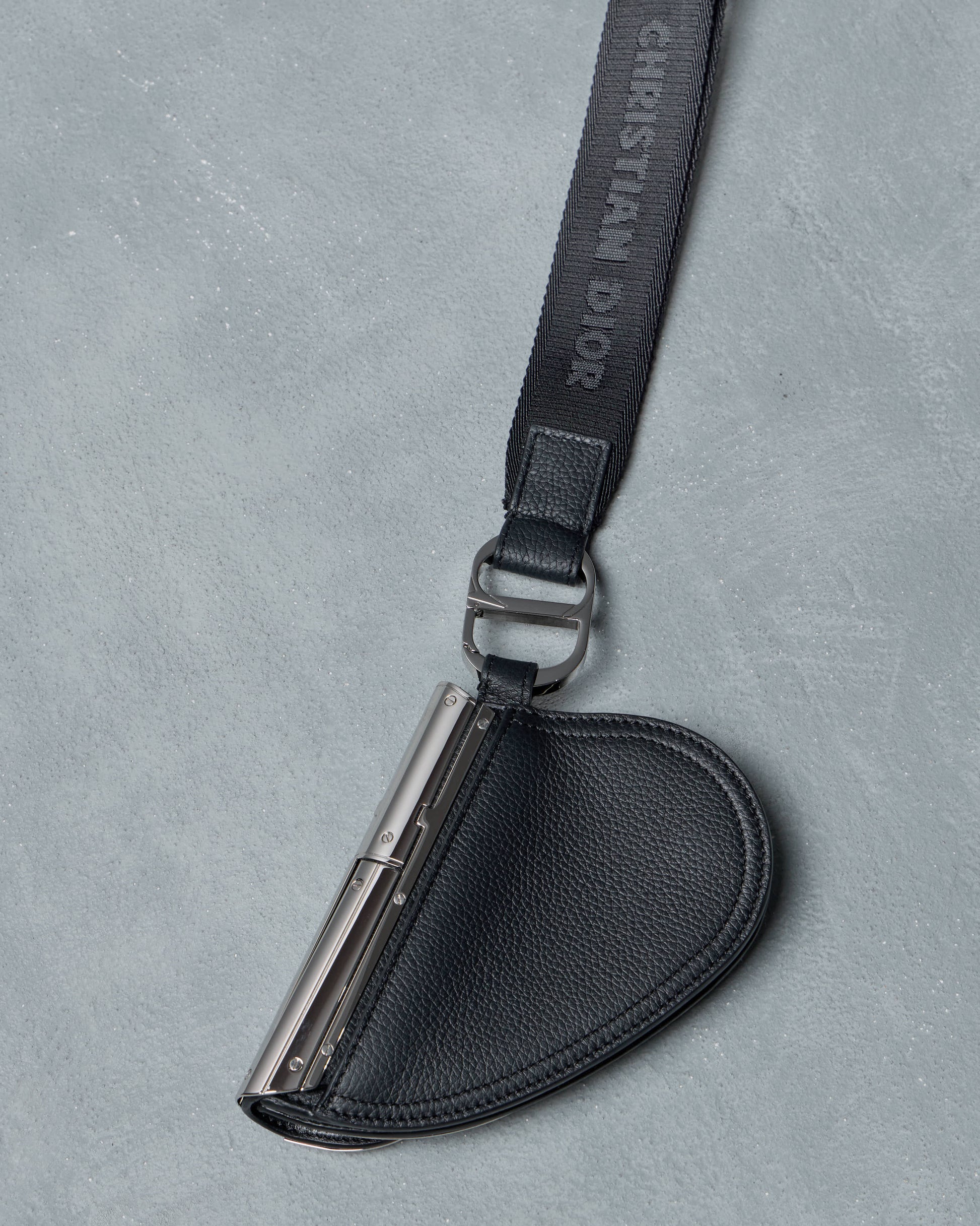 Black leather Dior case with a metallic clip on a gray background, featuring Christian Dior branding.
