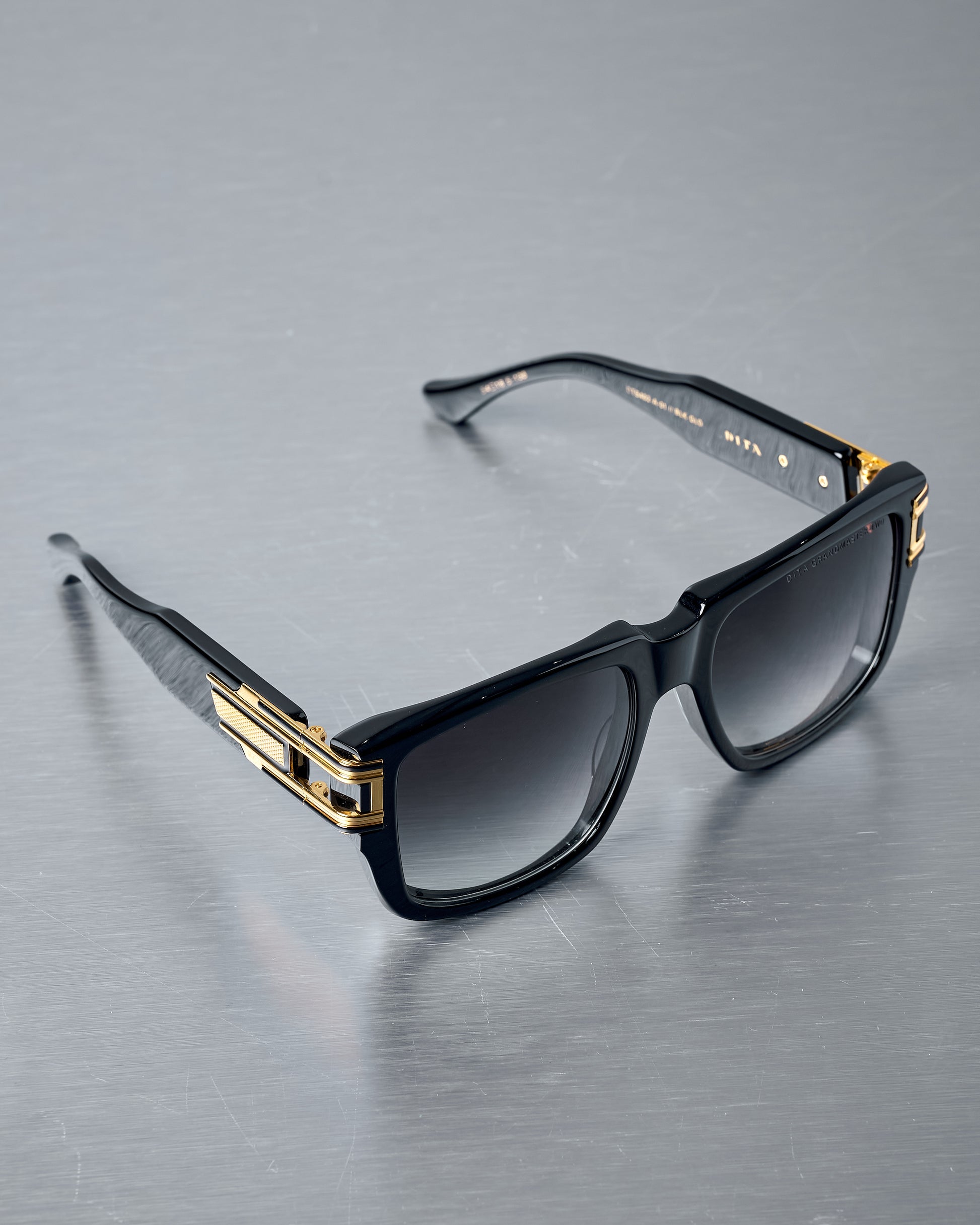 Black Dita sunglasses with gold accents on a reflective grey surface