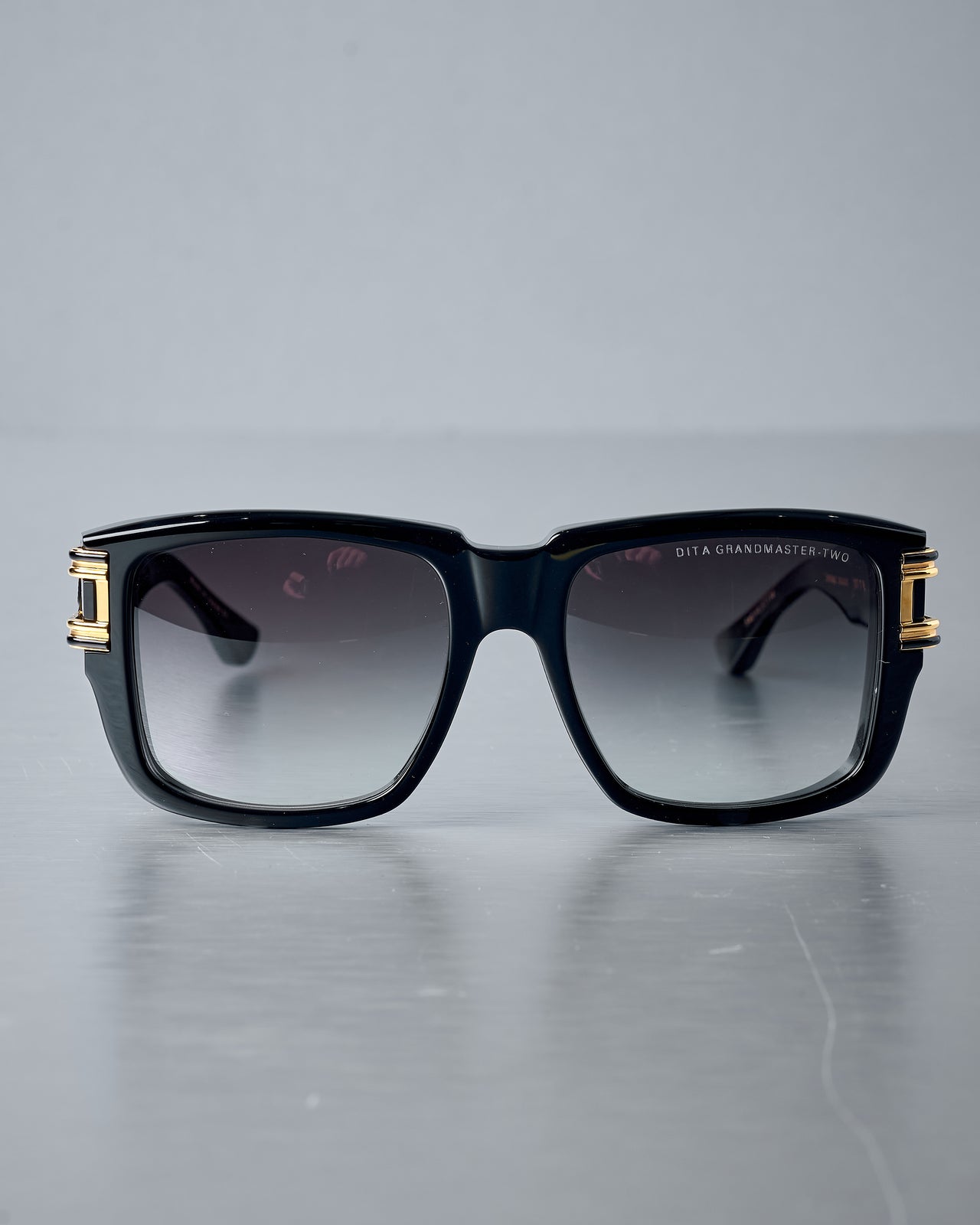 Black Dita sunglasses with gold accents on a reflective grey surface
