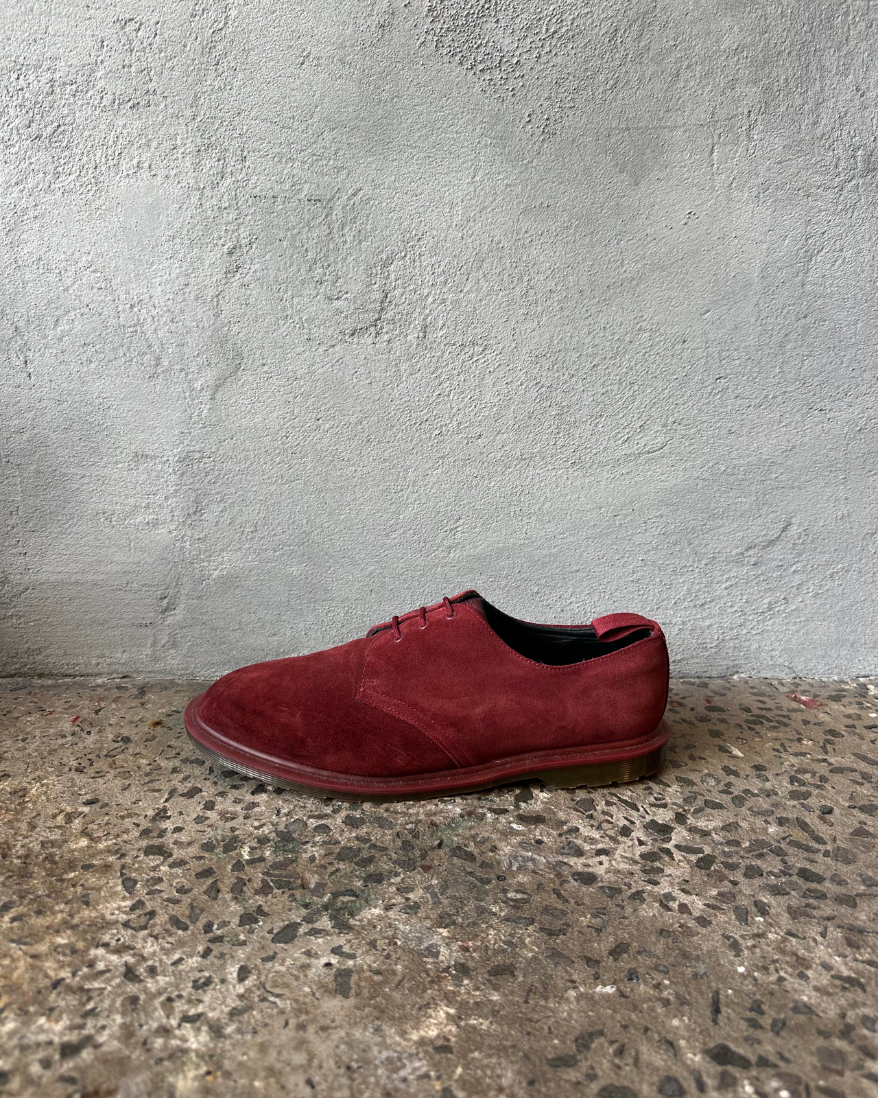 Red Doc Martens suede shoe on a textured concrete floor with a gray wall background