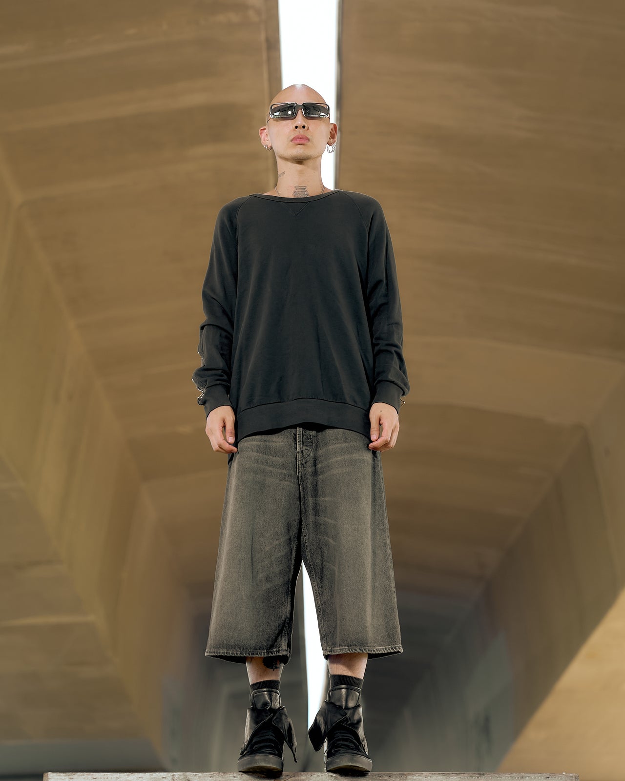 A person wearing a long black raglan Dries Van Noten sweater with a double-zip and a ribbed crewneck, paired with long beige shorts and black shoes, standing in front of a geometric-patterned wall.