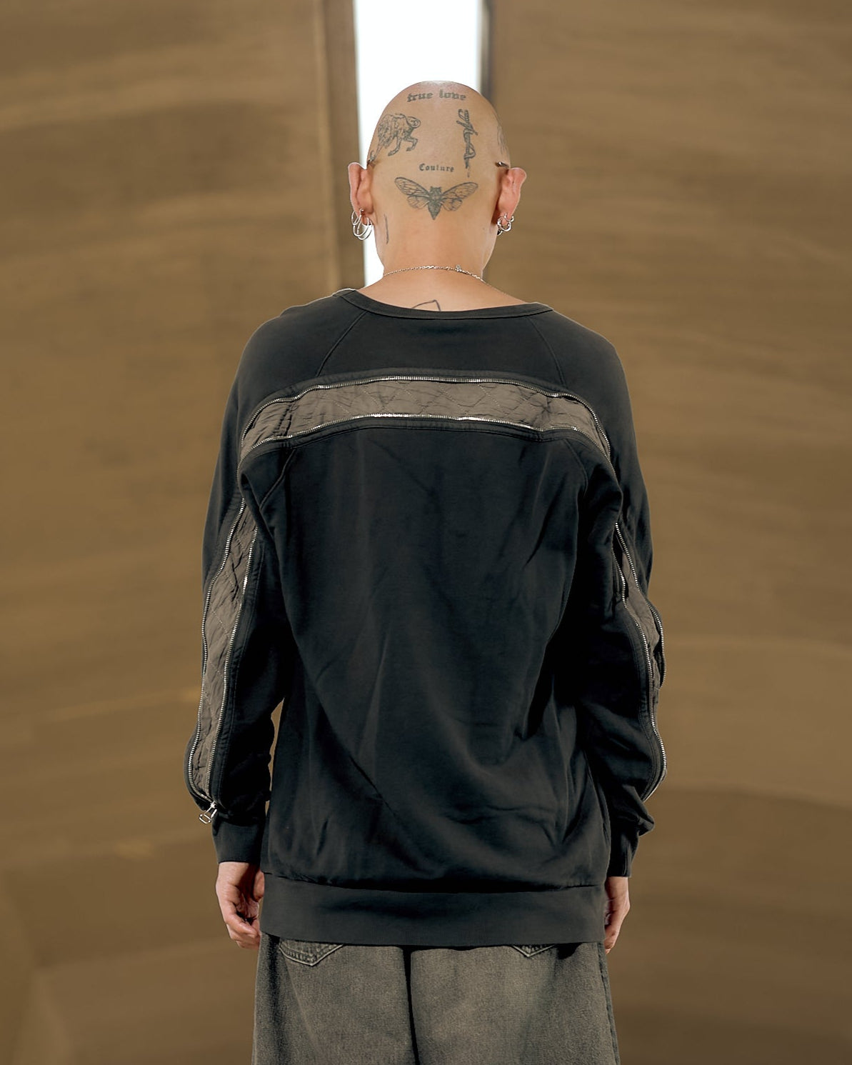 A person wearing a long black raglan Dries Van Noten sweater with a double-zip and a ribbed crewneck, paired with long beige shorts and black shoes, standing in front of a geometric-patterned wall.