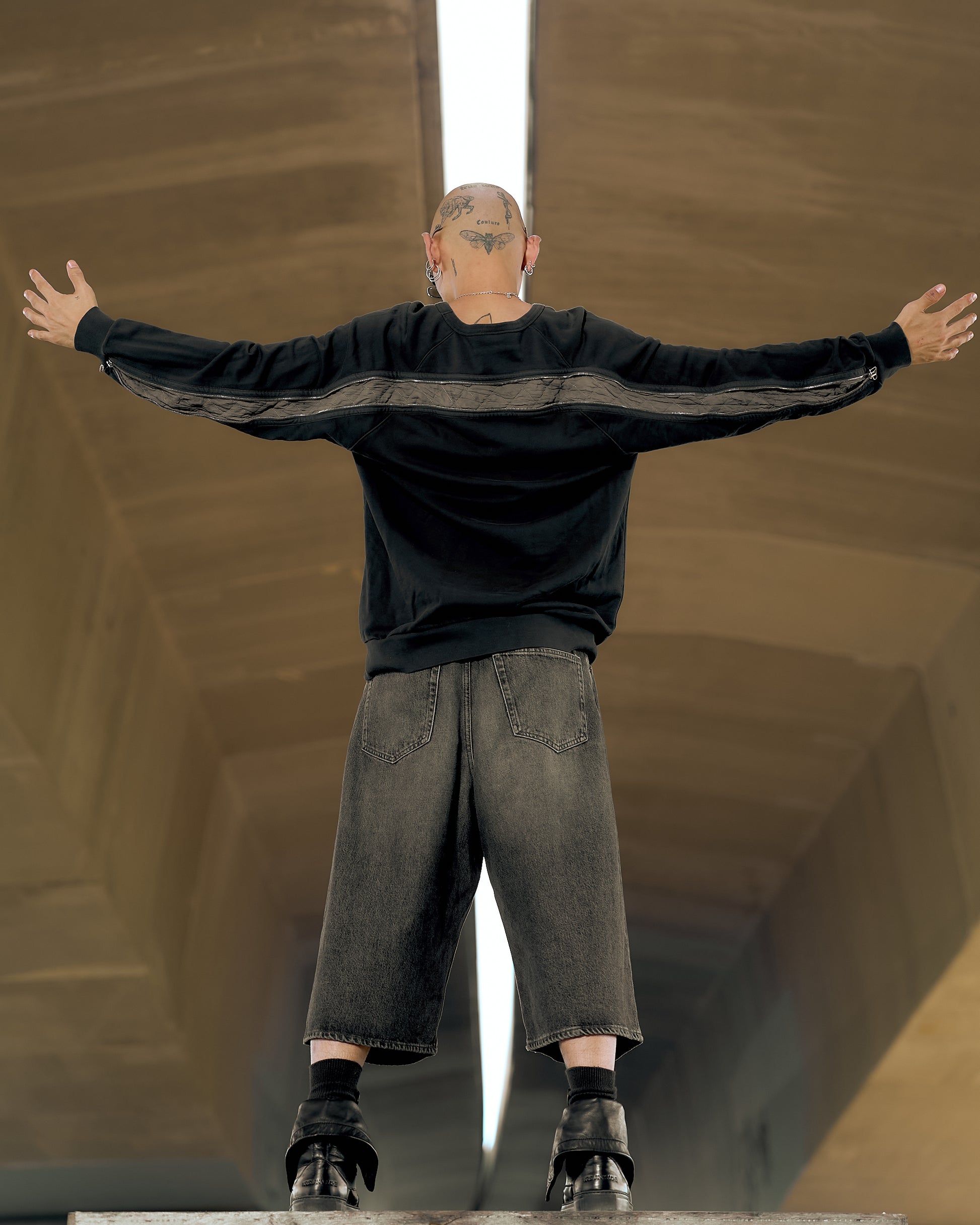 Person stands arms outstretched under a bridge, wearing a black Dries Van Noten sweatshirt and gray shorts. Bald, tattooed head visible. Urban, defiant tone.