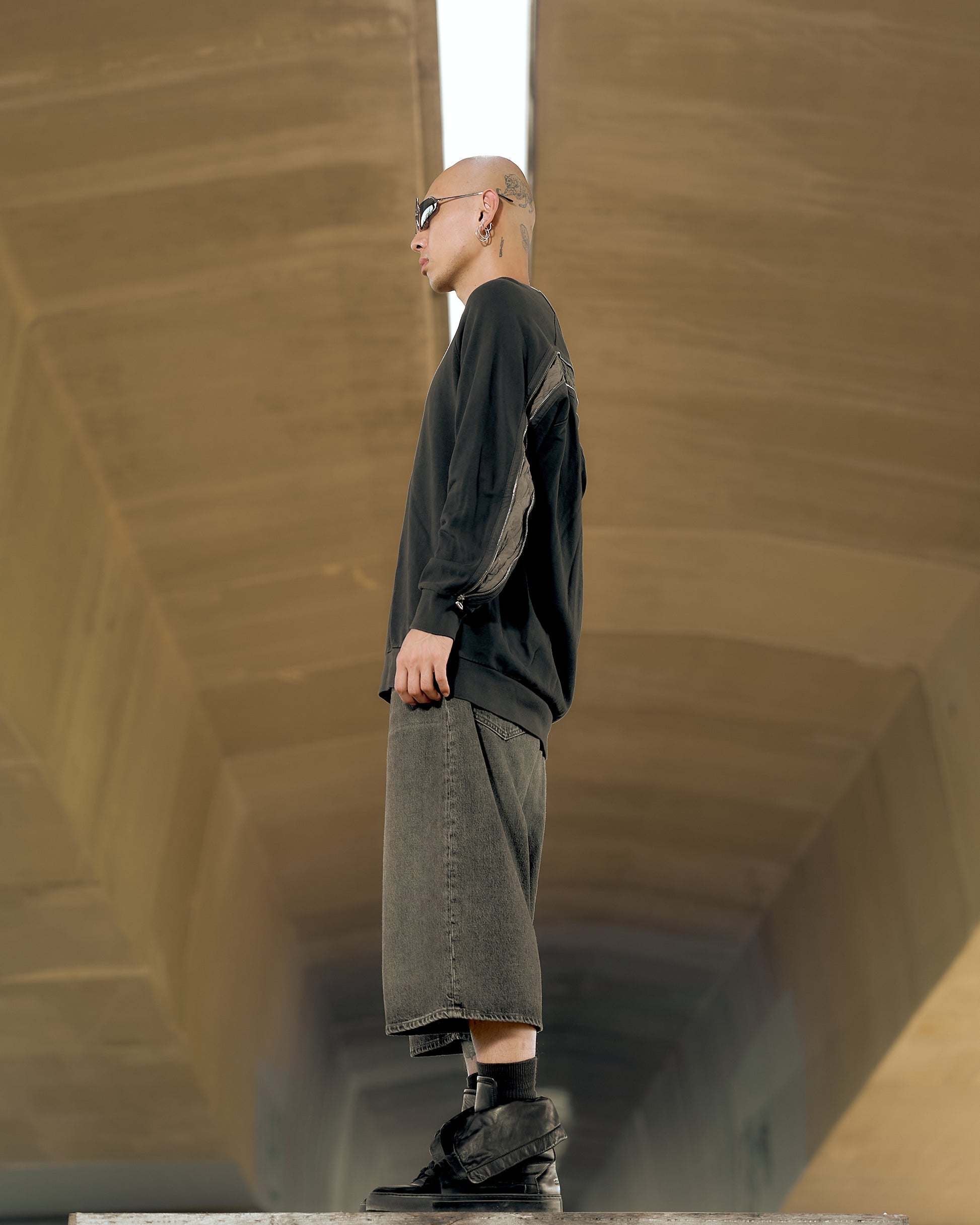 A person stands confidently in an urban setting wearing sunglasses, a black Dries Van Noten sweater with a zipper detail, and loose, gray shorts. The background features an expansive, modern tunnel ceiling.