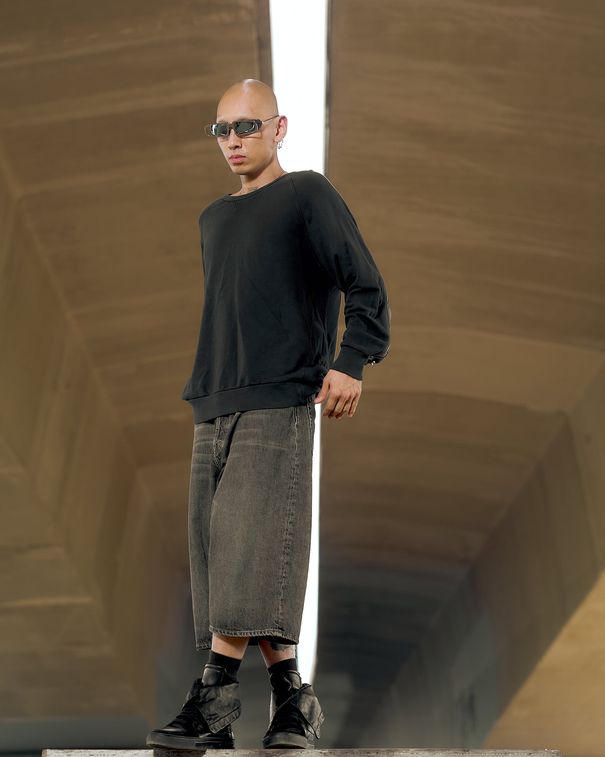 A person stands confidently in an urban setting wearing sunglasses, a black Dries Van Noten sweater with a zipper detail, and loose, gray shorts. The background features an expansive, modern tunnel ceiling.