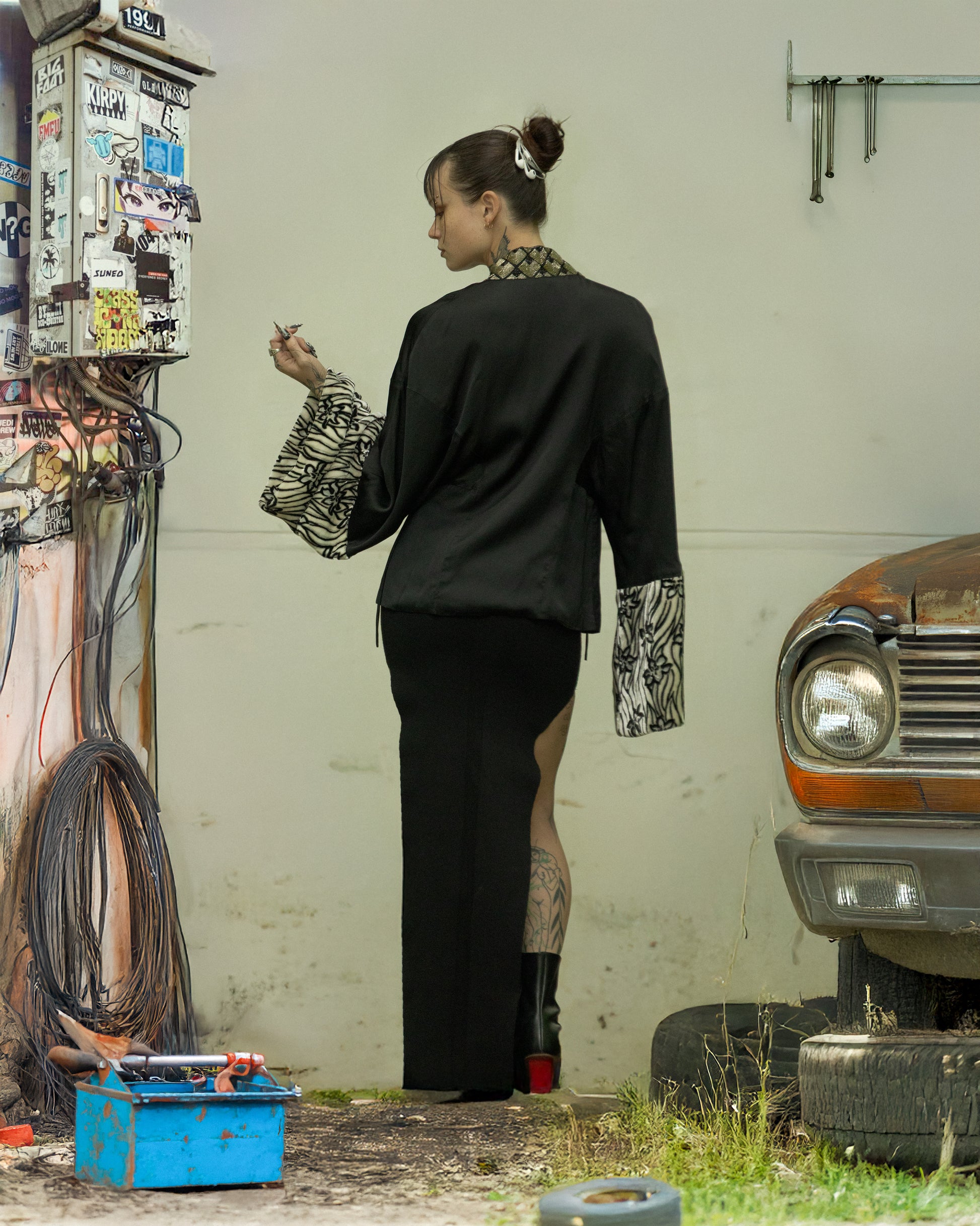 Person standing in a garage with a vintage car and colorful wall in the background. They are wearing a Dries Van Noten beaded kimono top.