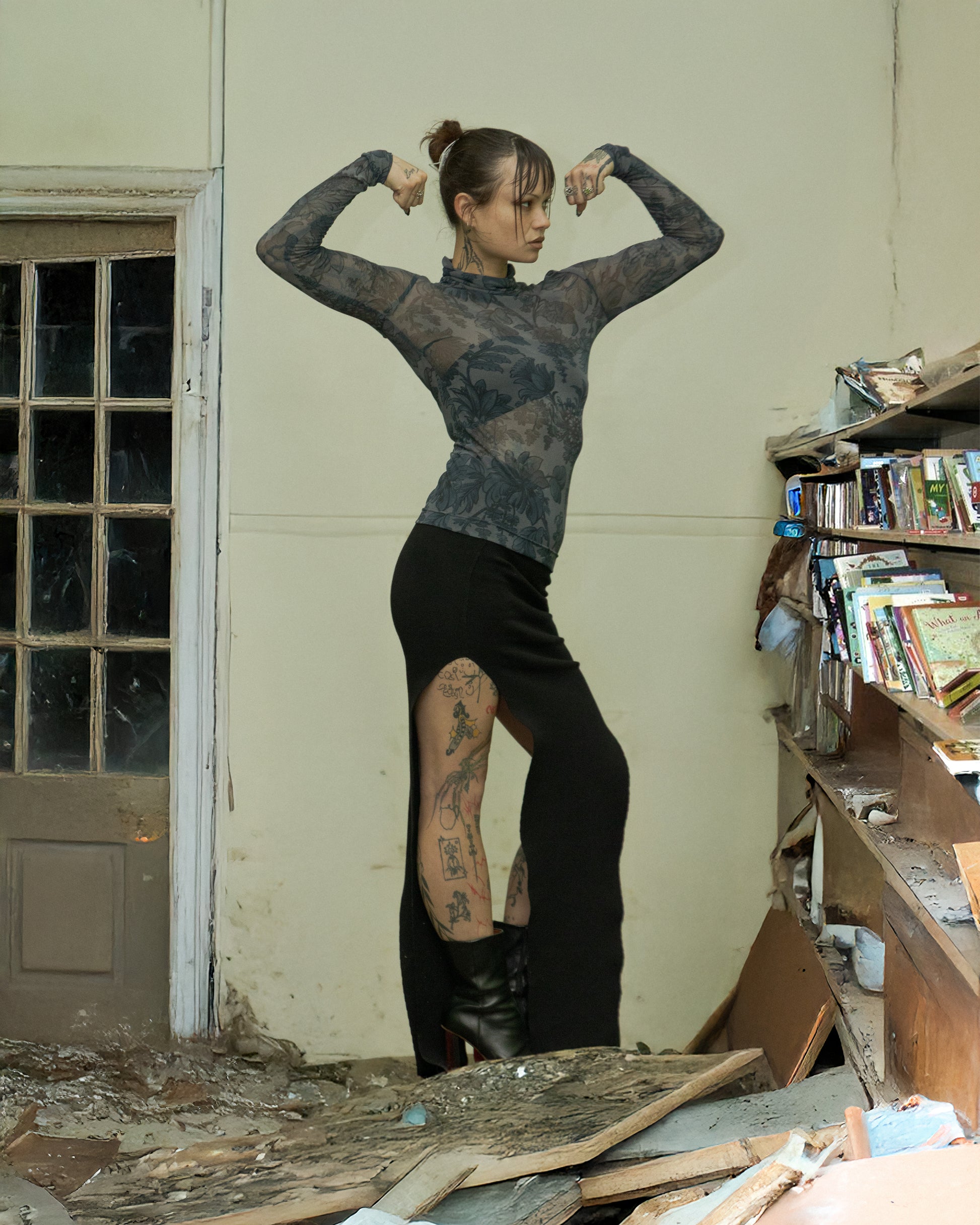 Person flexing muscles in a room with books and debris wearing a sheer flora Dries Van Noten top.