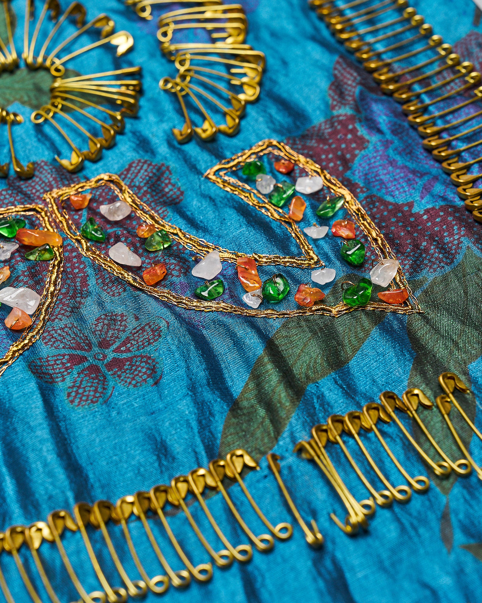 Close-up of a vibrant blue Dries Van Noten fabric adorned with safety pins, gold embroidery, and multicoloured stones, arranged in floral and abstract patterns.