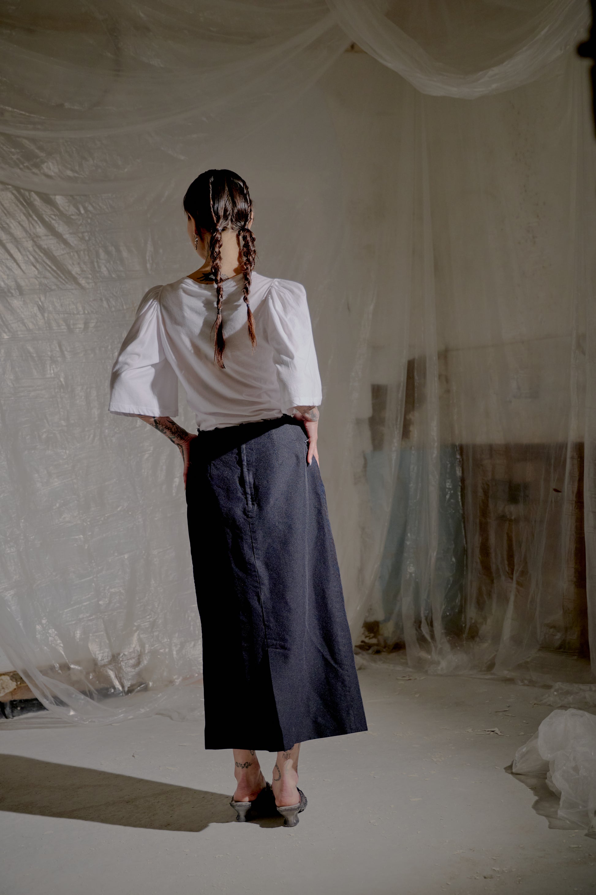 Person wearing a white Dries Van Noten shirt and dark pants standing in a room with white plastic sheeting.