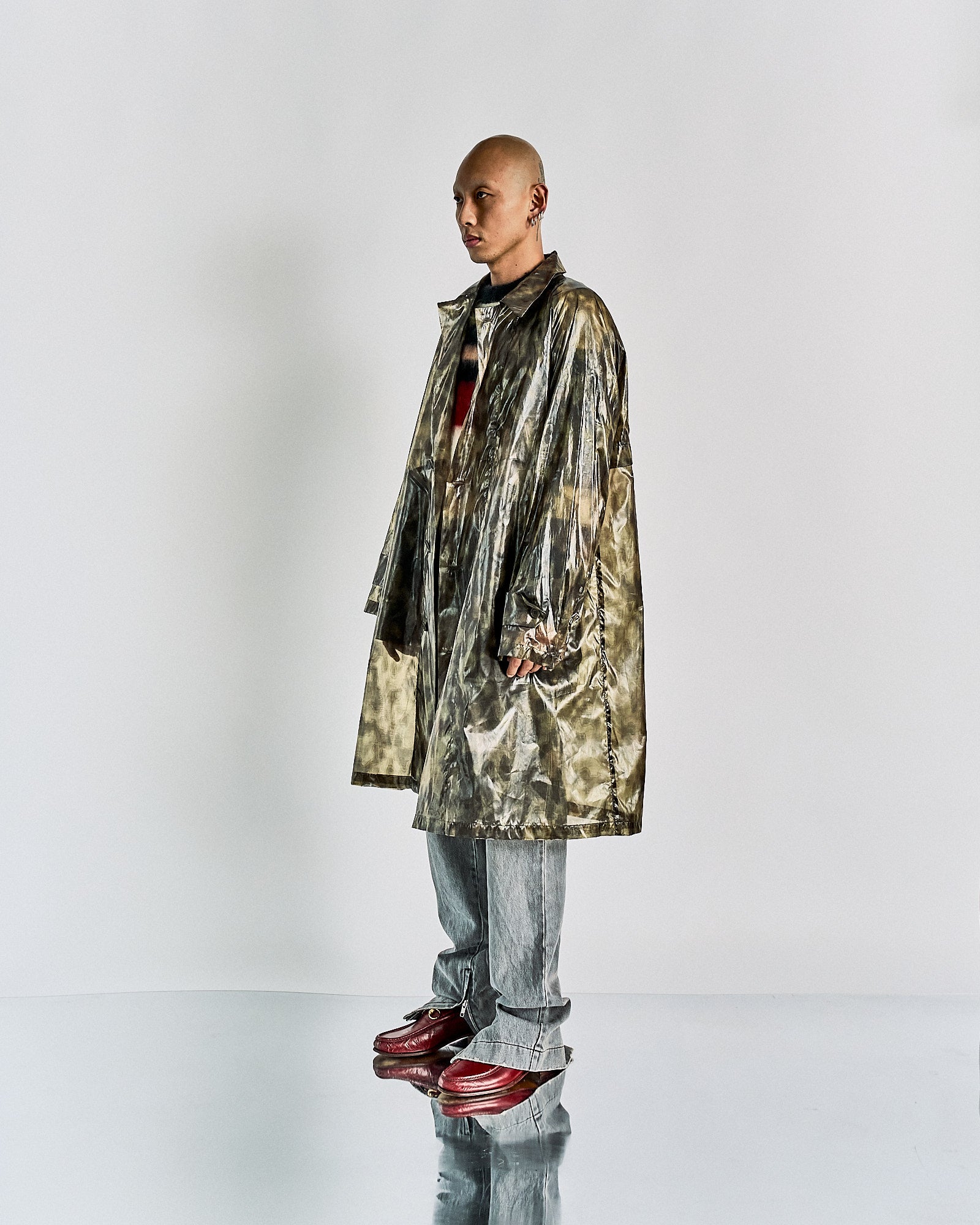 A person stands confidently on a reflective surface wearing a shiny, oversized metallic Dries Van Noten coat, grey pants, and red shoes. The expression is serious, conveying a futuristic vibe.