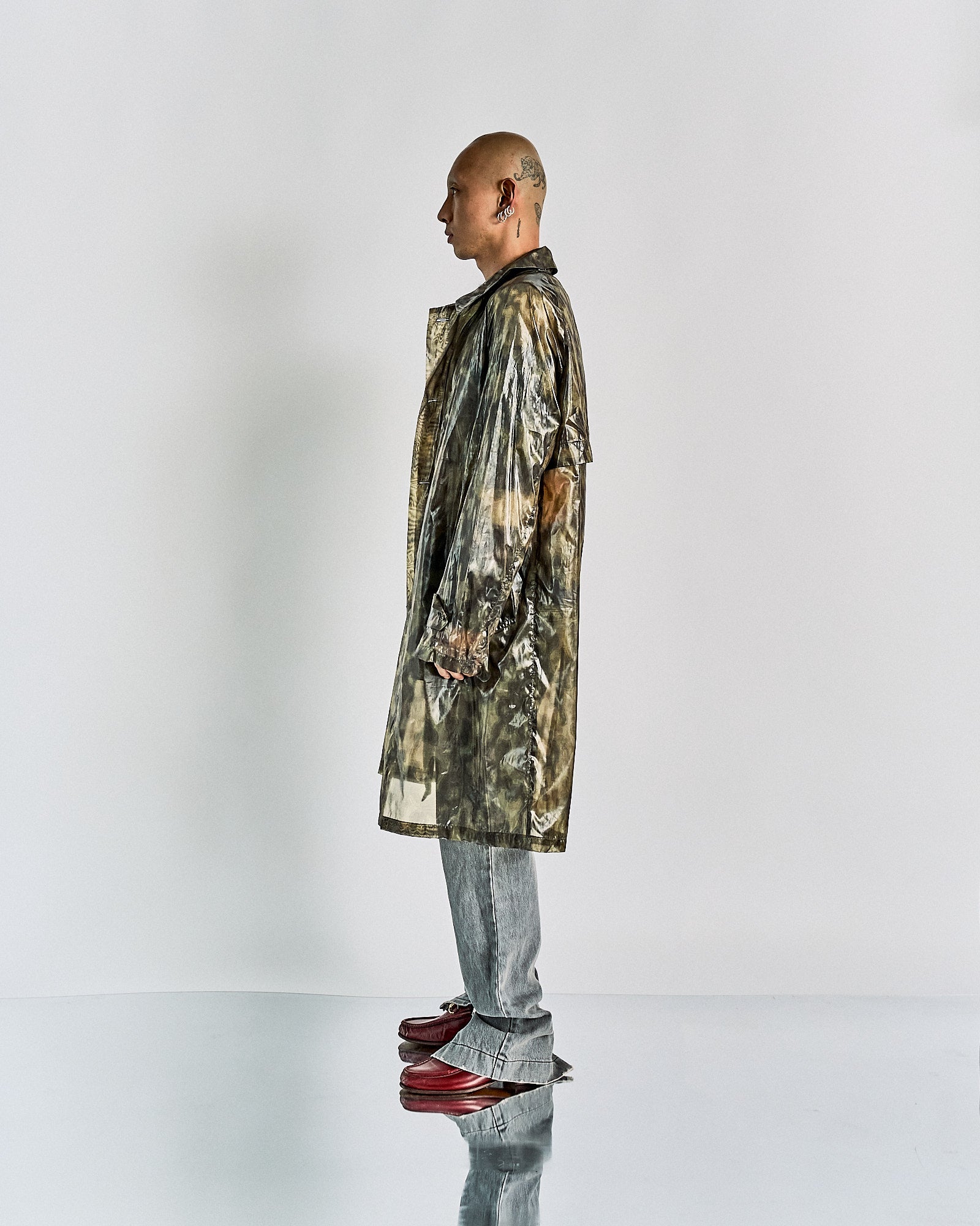 A person stands in profile against a plain background, wearing a long, shiny, patterned Dries Van Noten coat with gray jeans and red shoes, conveying a chic, modern style.
