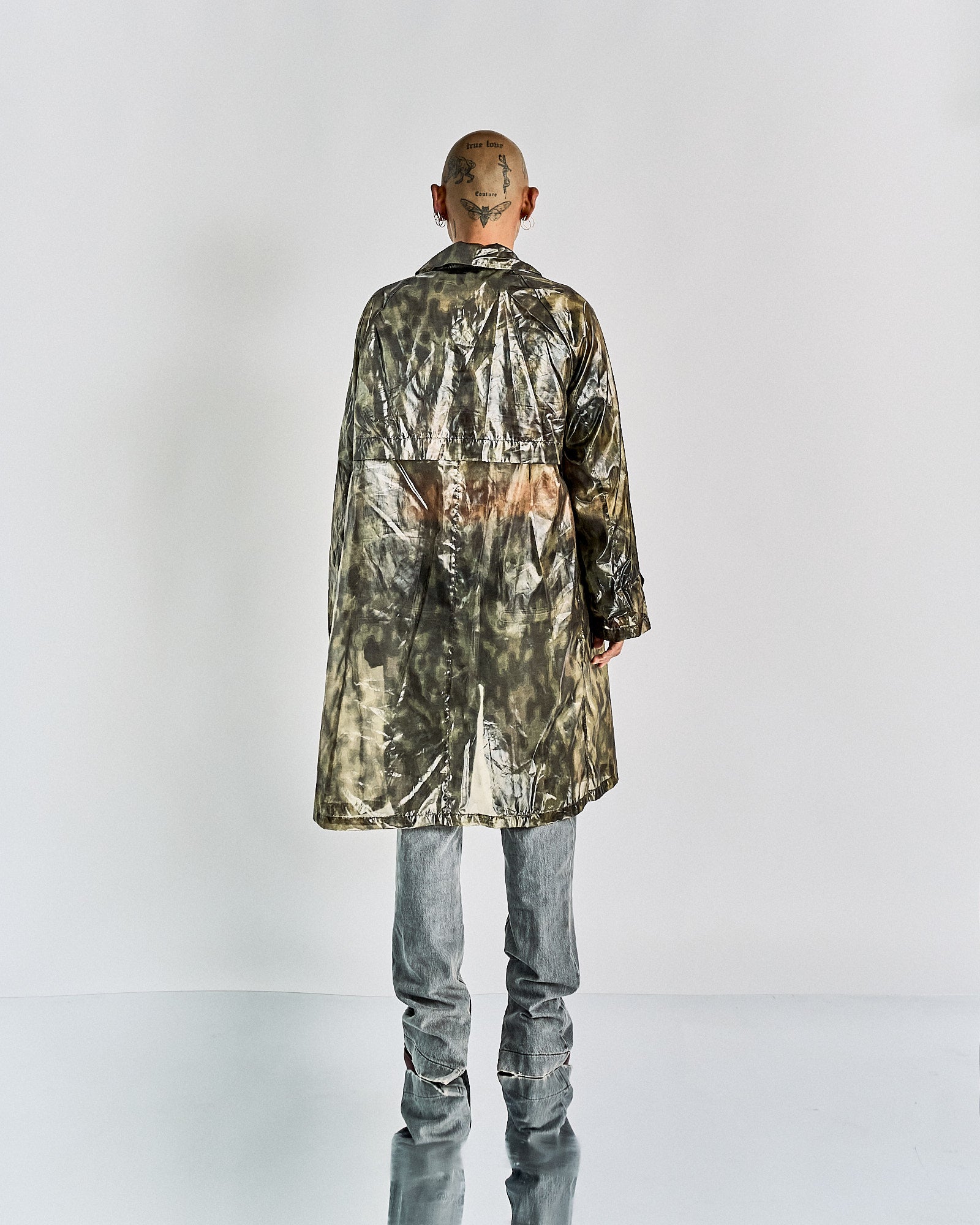 A person stands with their back to the camera, wearing a long, reflective, tie-dye Dries Van Noten coat and gray pants. The setting is minimal, emphasizing the outfit's texture.